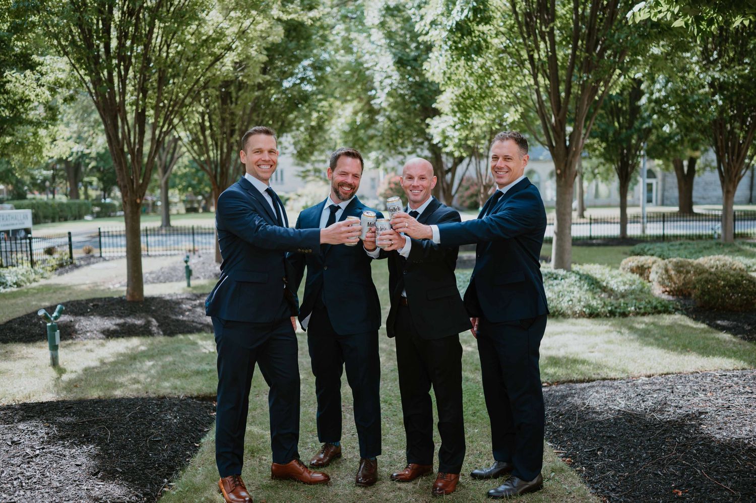 Four groomsmen in navy suits toast together outdoors in a tree-lined park before wedding ceremony.