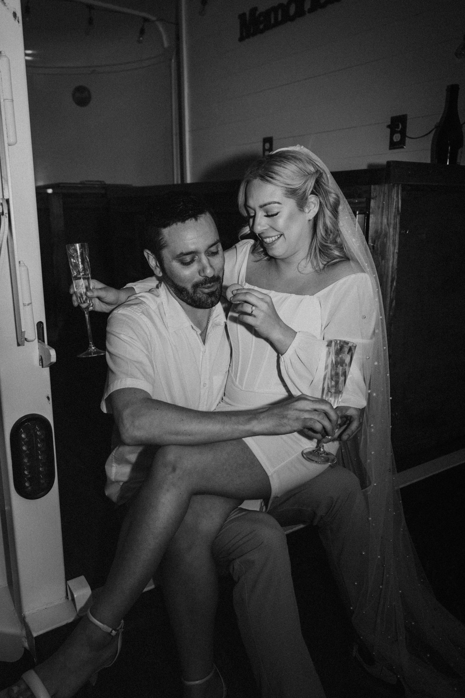 Newlyweds toast champagne glasses while sitting together in a cozy van in black and white.