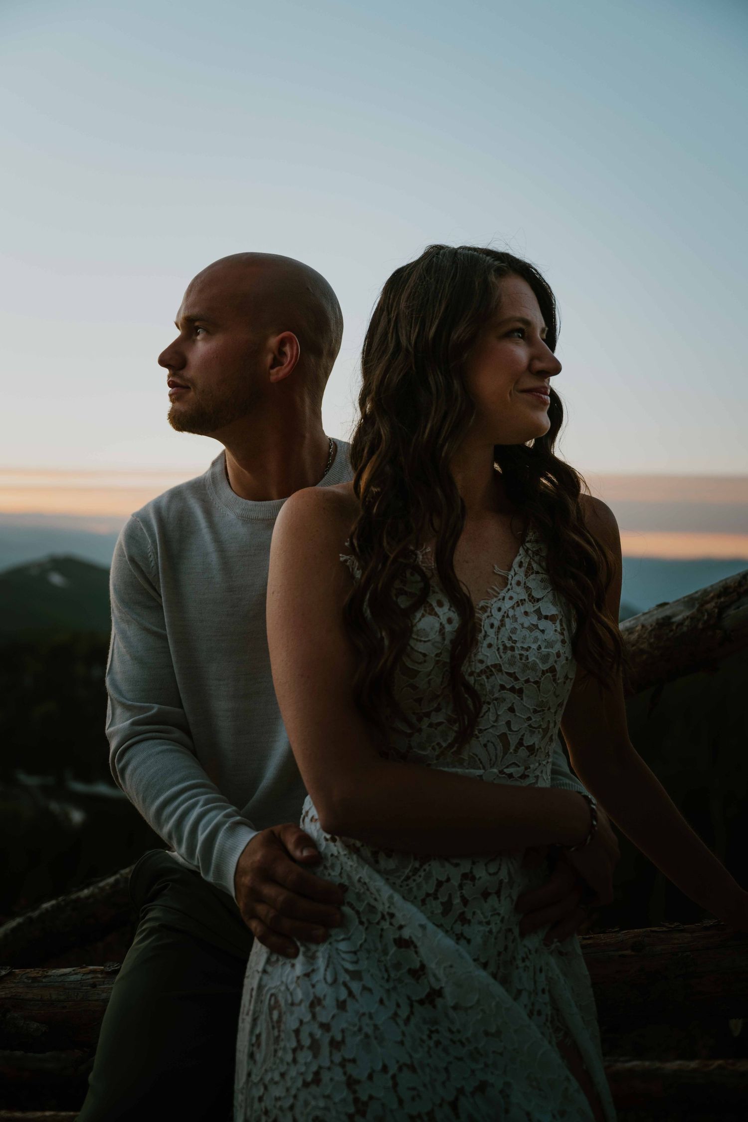 Couple poses back-to-back at sunset with mountain views, woman in lace dress, man in gray sweater.