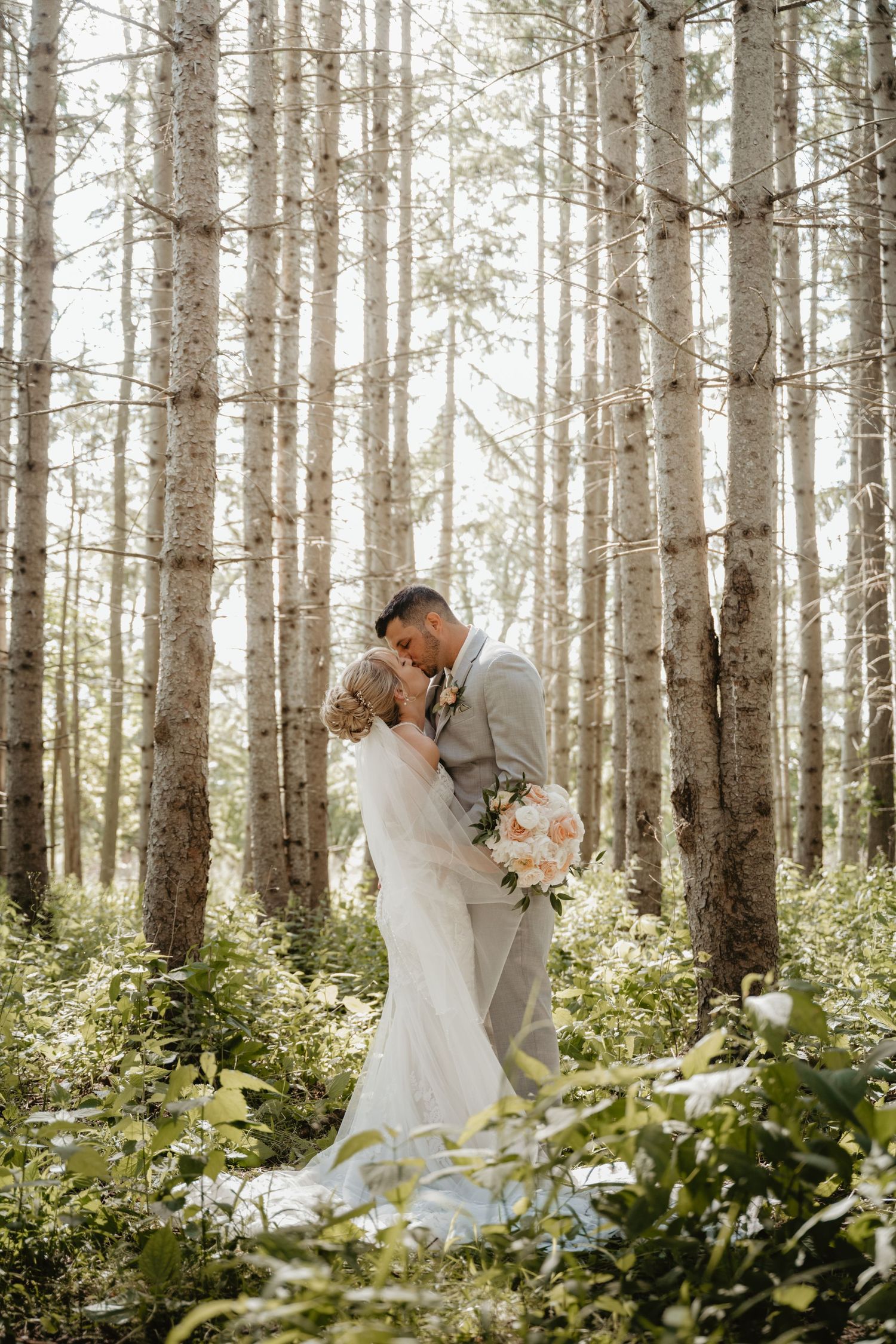 Bride and groom share a romantic kiss surrounded by tall pine trees in a sunlit forest wedding.
