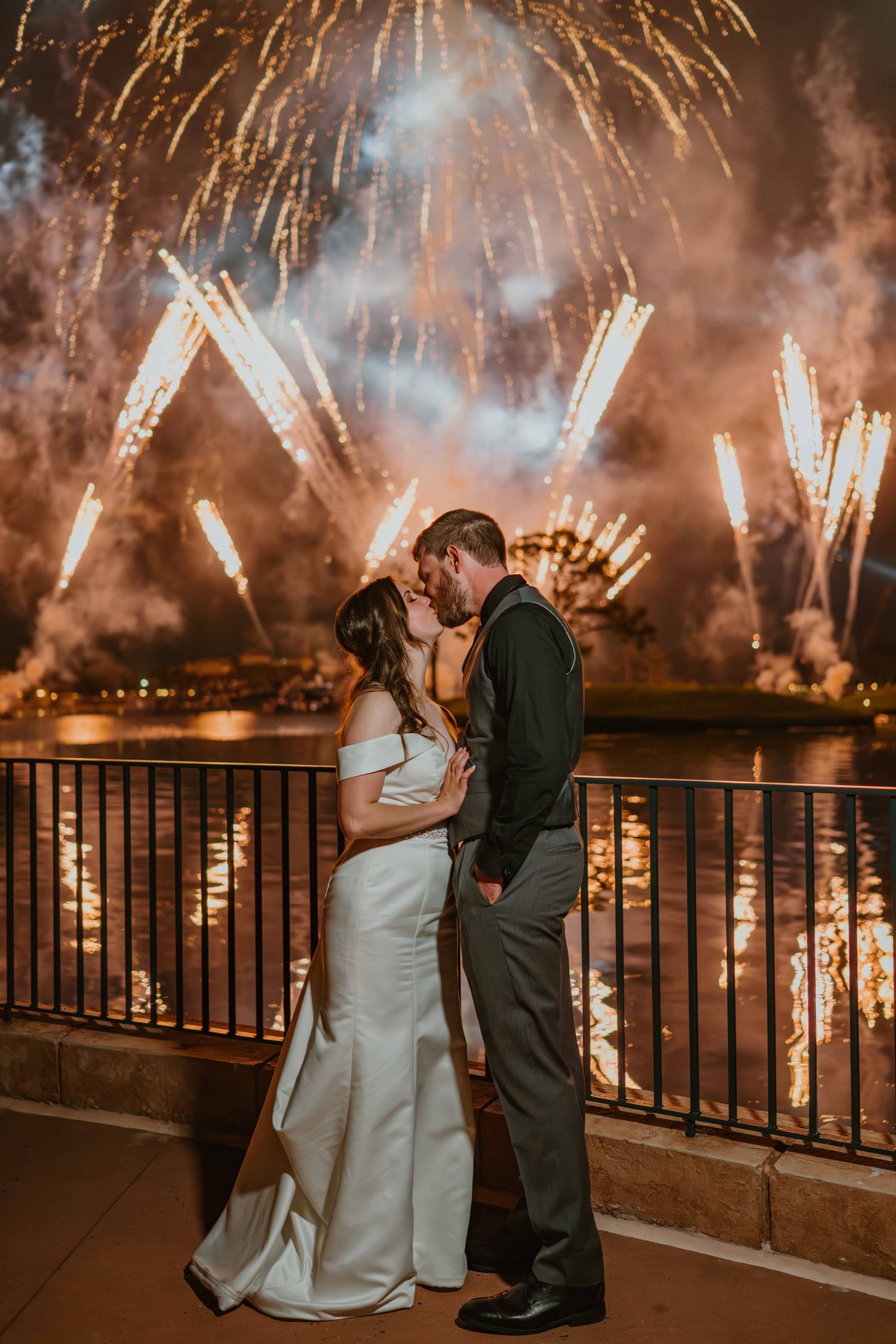 Wedding couple kissing on waterfront balcony with spectacular colorful fireworks display lighting night sky.
