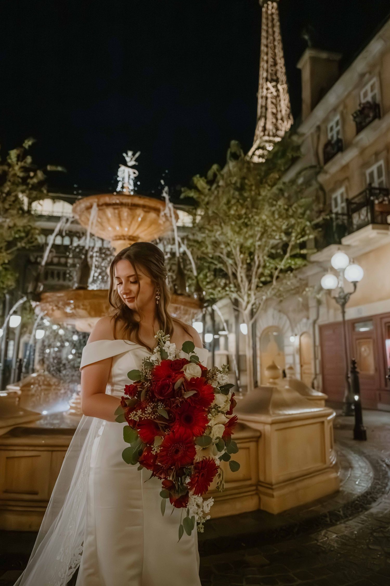 Bride in off-shoulder gown holding red rose bouquet near fountain with lit Eiffel Tower.