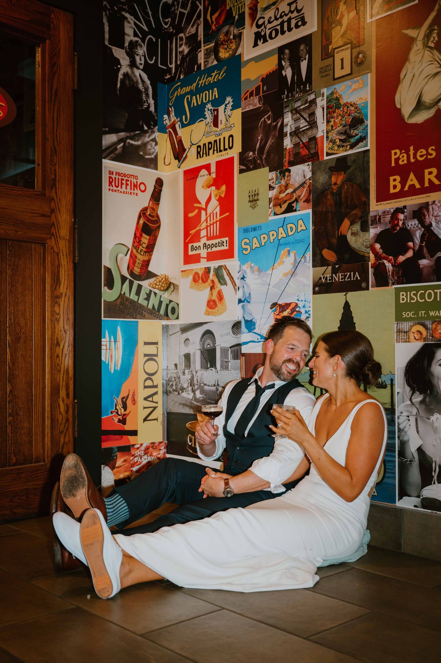 Bride and groom sit laughing together on floor against vintage Italian travel poster wall at wedding reception.