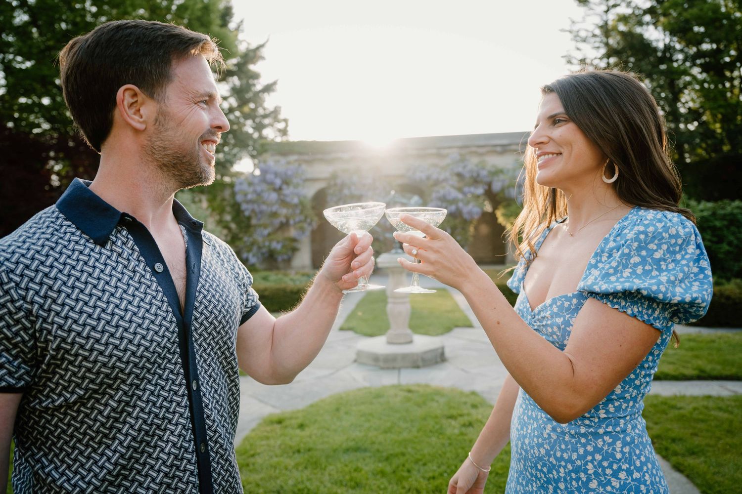 Couple toasts martini glasses outdoors in sunlit garden during romantic engagement photo session.