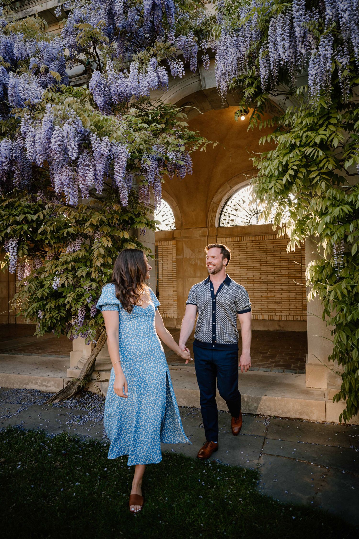 Couple holds hands under blooming purple wisteria at a historic stone garden archway in golden hour light.