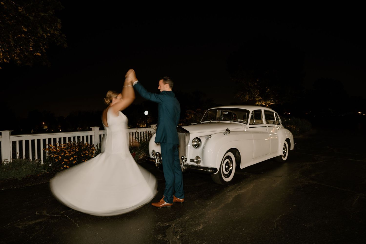 Groom twirling bride in a flowing white gown beside a vintage white car on their wedding night.