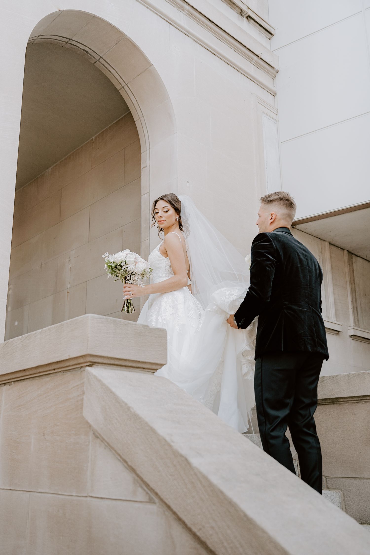 Bride and groom holding hands while ascending stone steps at classic architectural outdoor wedding venue.