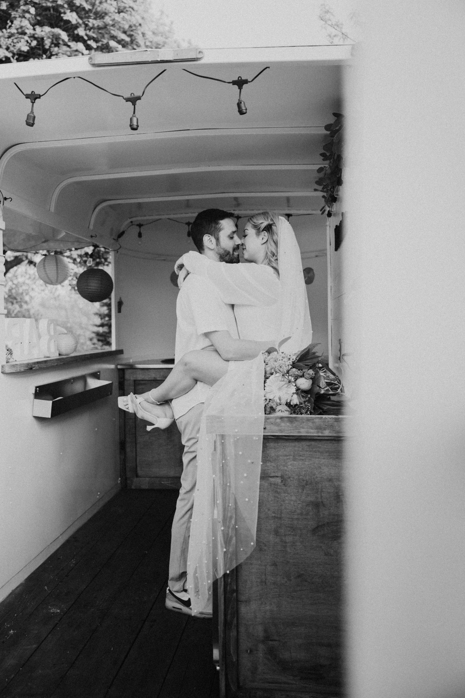 Black and white photo of groom lifting bride for a kiss inside a rustic mobile bar truck.