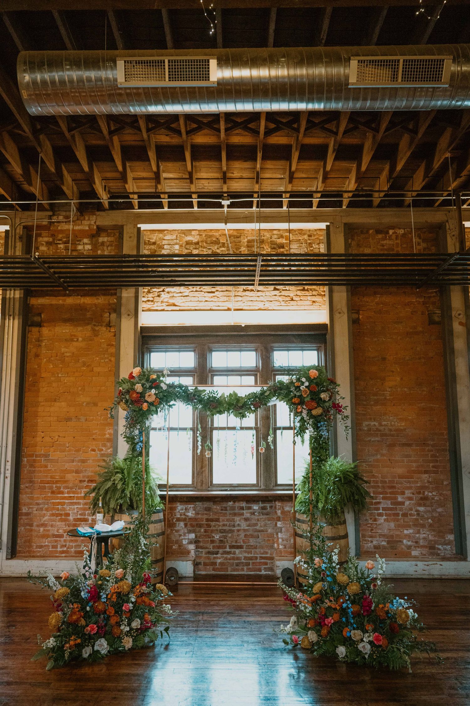 Vibrant floral wedding ceremony arch with greenery displayed in a rustic brick loft venue with large windows.