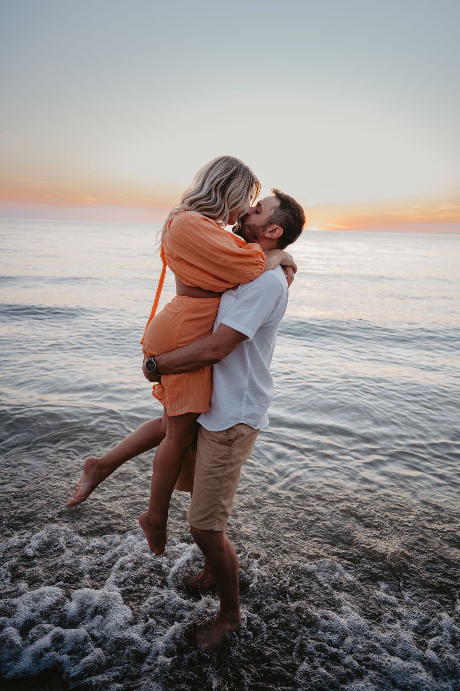 Man lifts woman in orange dress as they kiss at the water's edge during a golden sunset beach session.