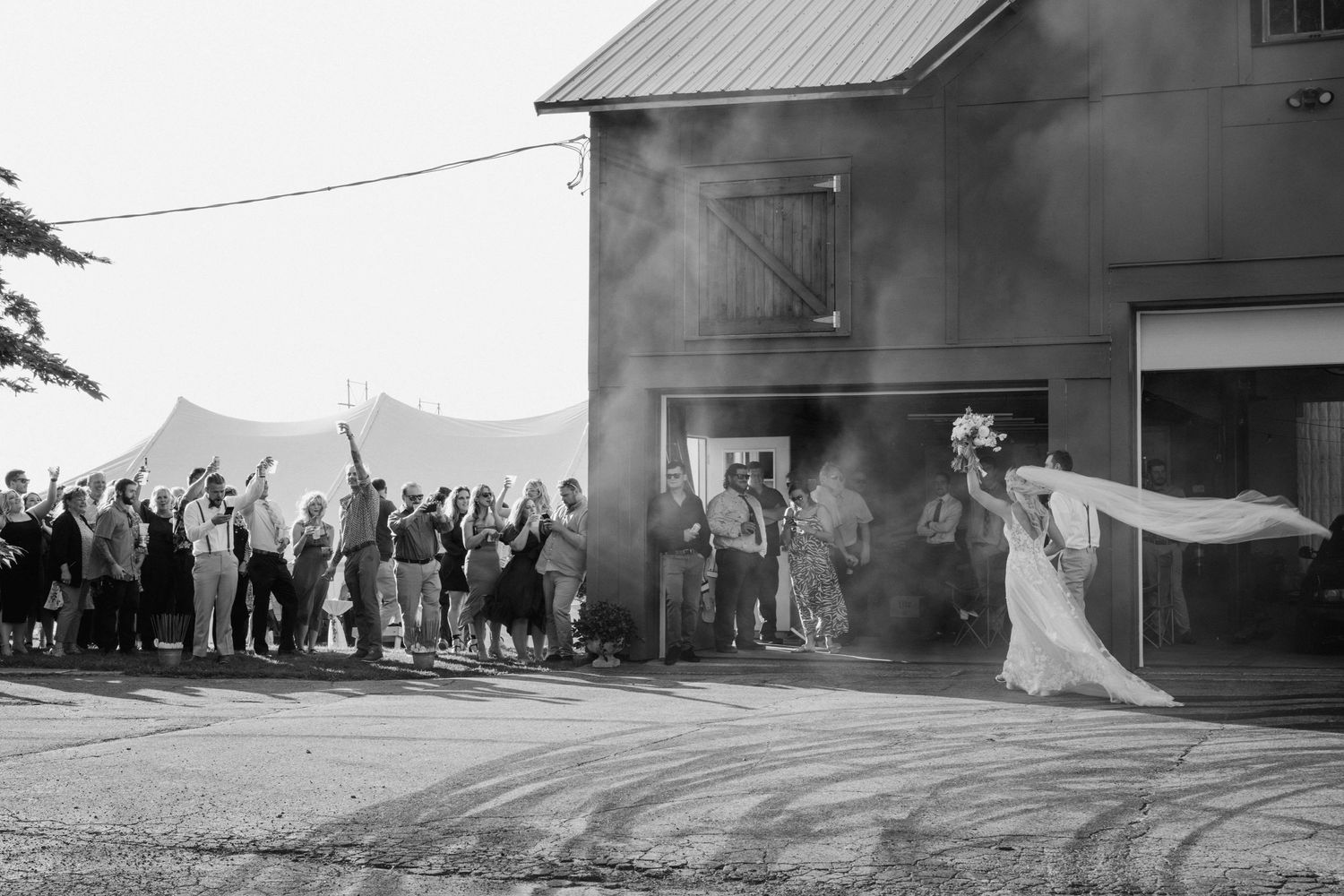 Black and white photo of wedding guests cheering as bride exits barn amid smoke and celebration.