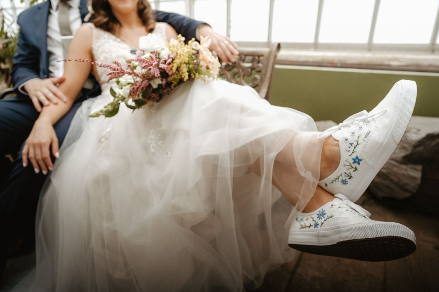 Bride showing custom embroidered white sneakers with colorful wedding bouquet on lap.
