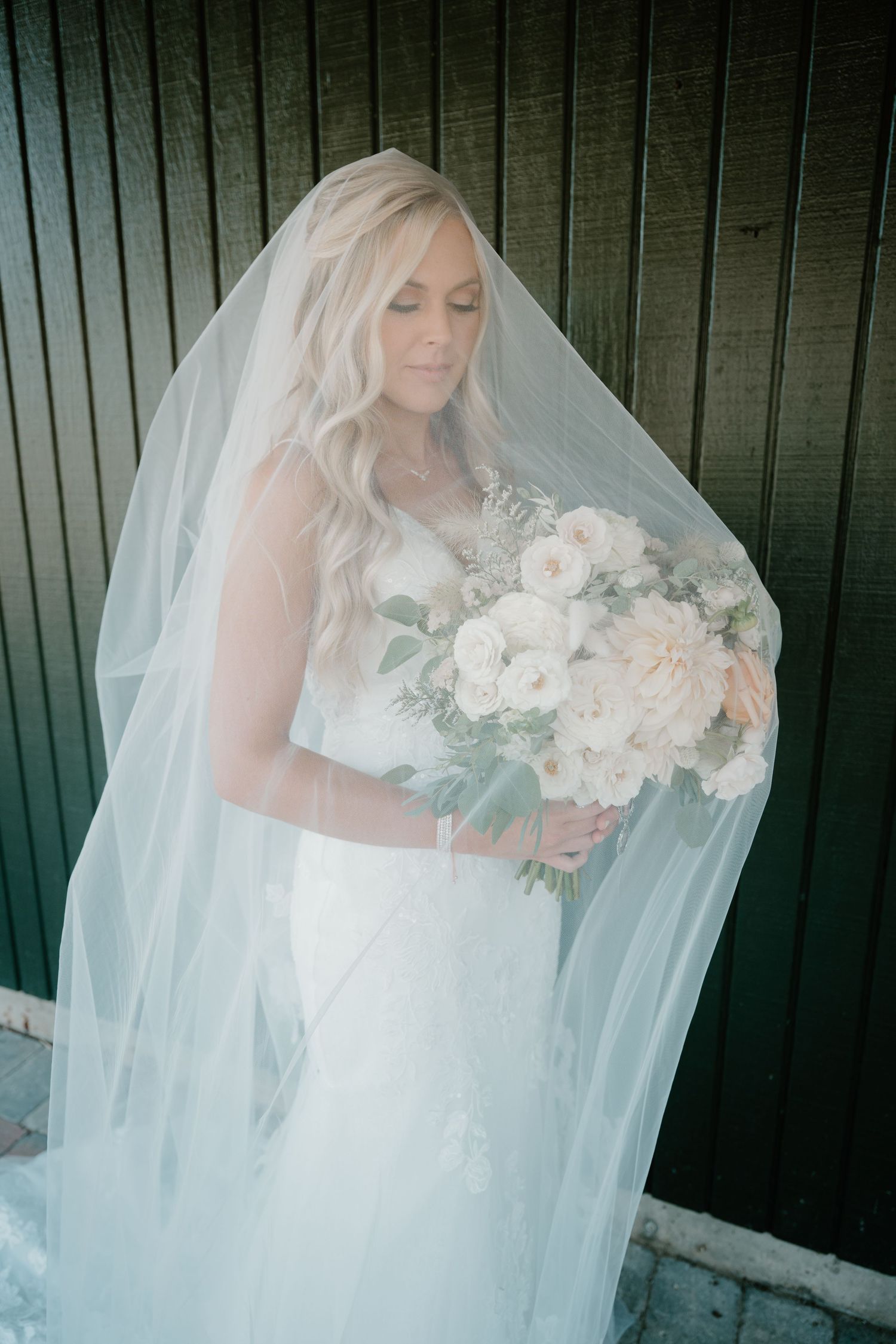 Bride in lace gown holding white floral bouquet, draped in flowing veil against dark wall.