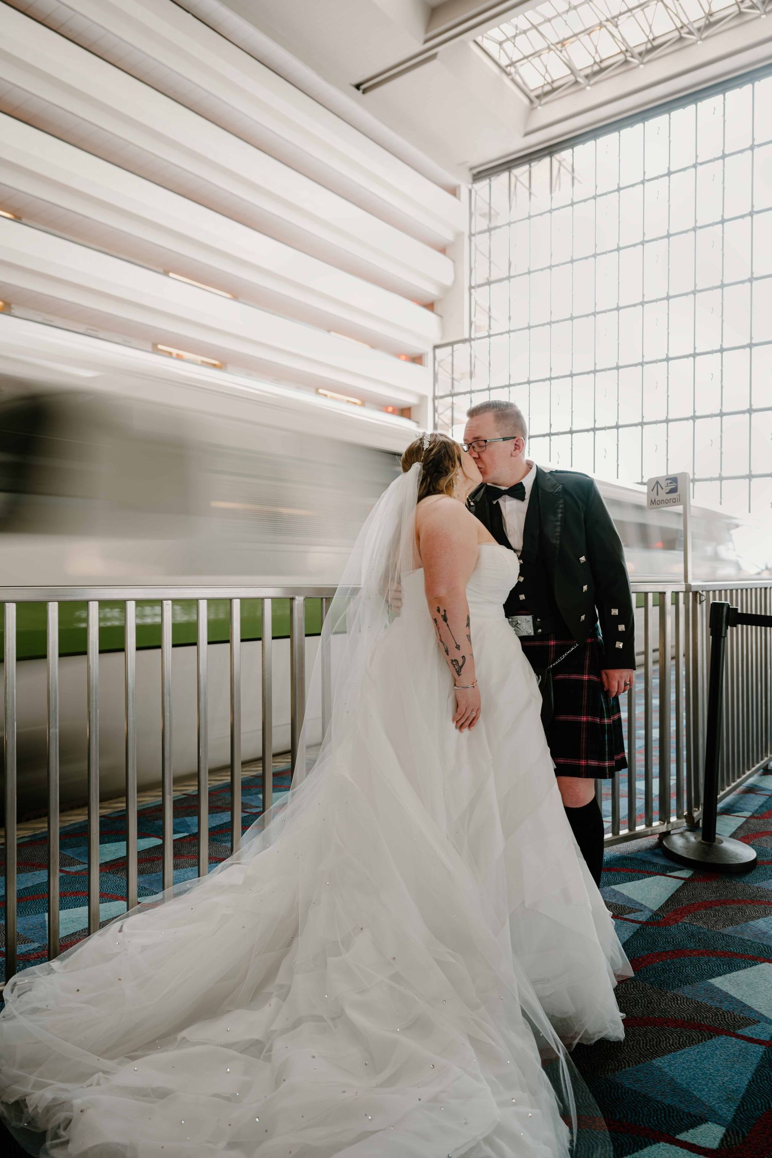 Bride in white gown and groom in Scottish kilt sharing a kiss inside Disney resort with monorail passing.