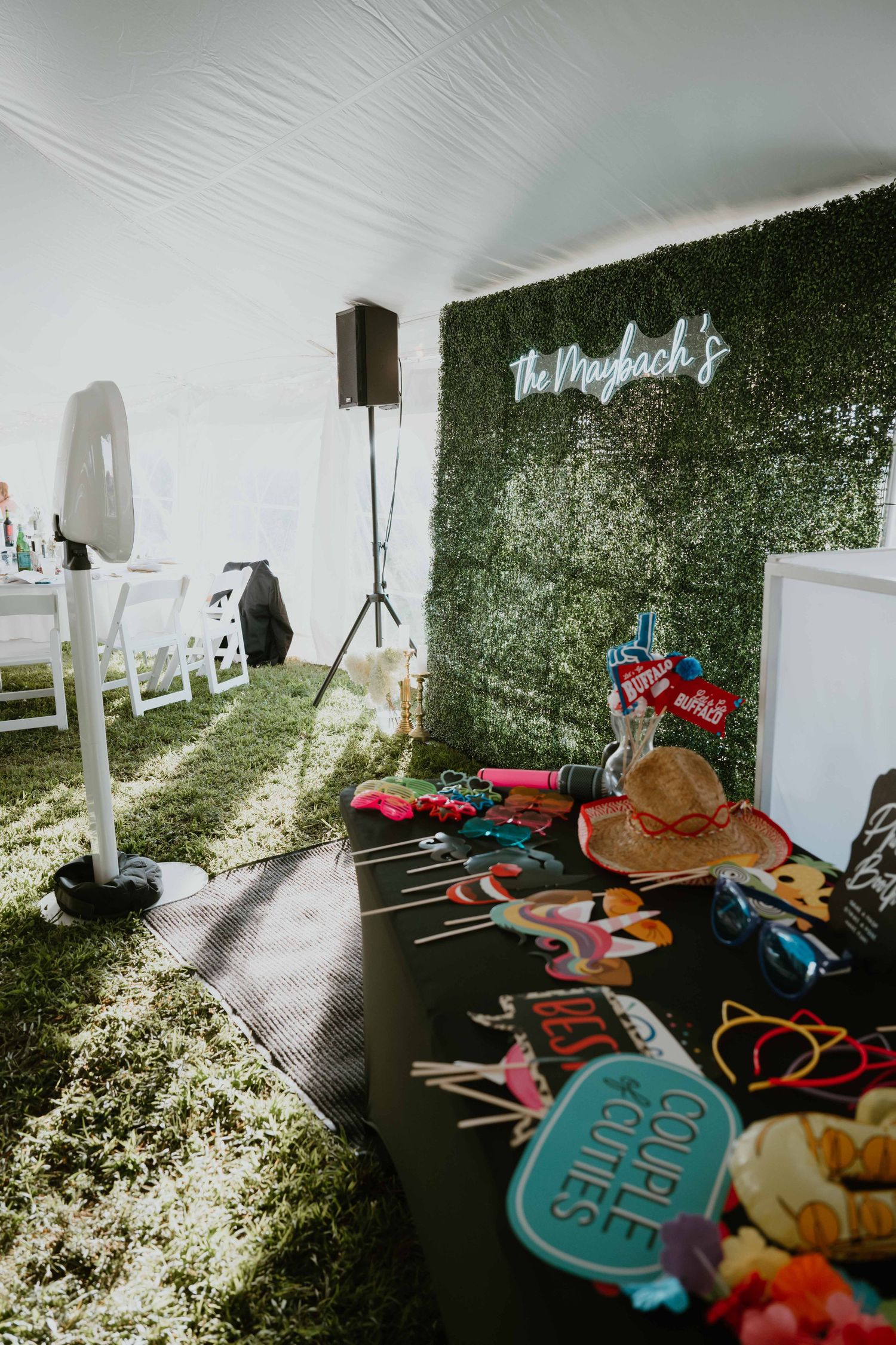 Wedding photo booth setup with green hedge wall, neon sign, and fun props under a white tent.