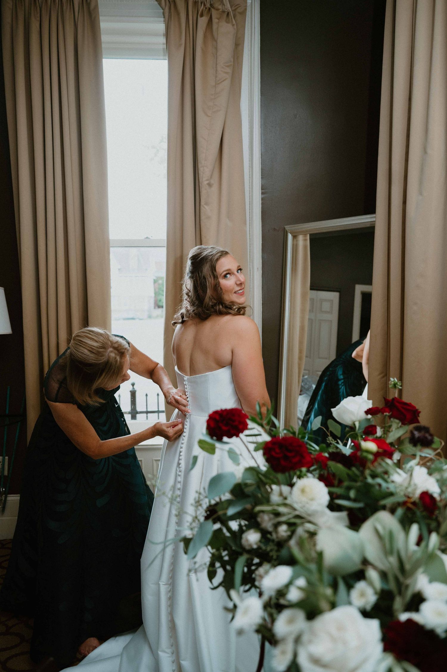 Bride smiling as assistant fastens wedding gown near window with red and white floral arrangements.