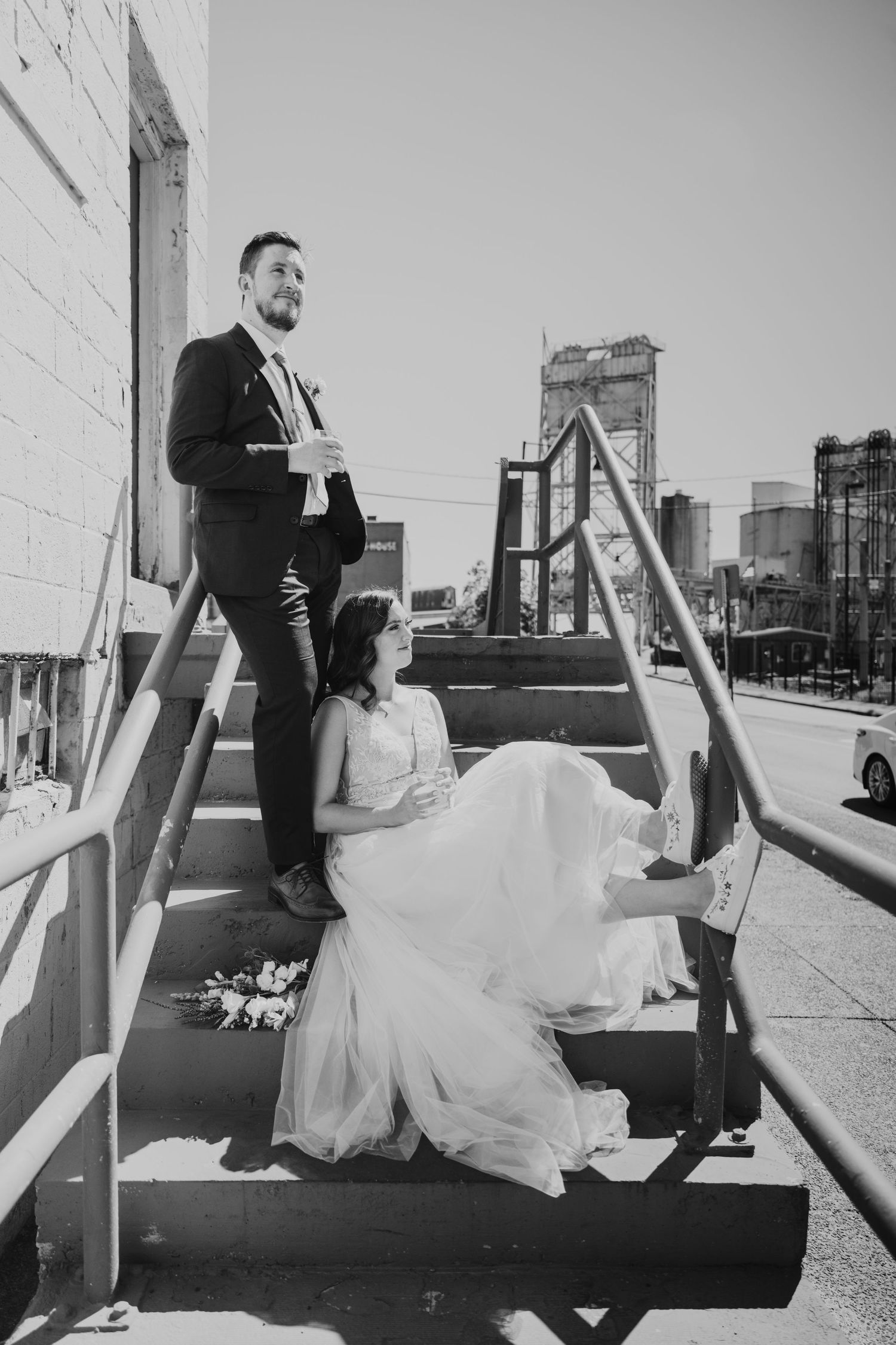Black and white photo of bride and groom posing on outdoor metal stairs with city skyline in background.