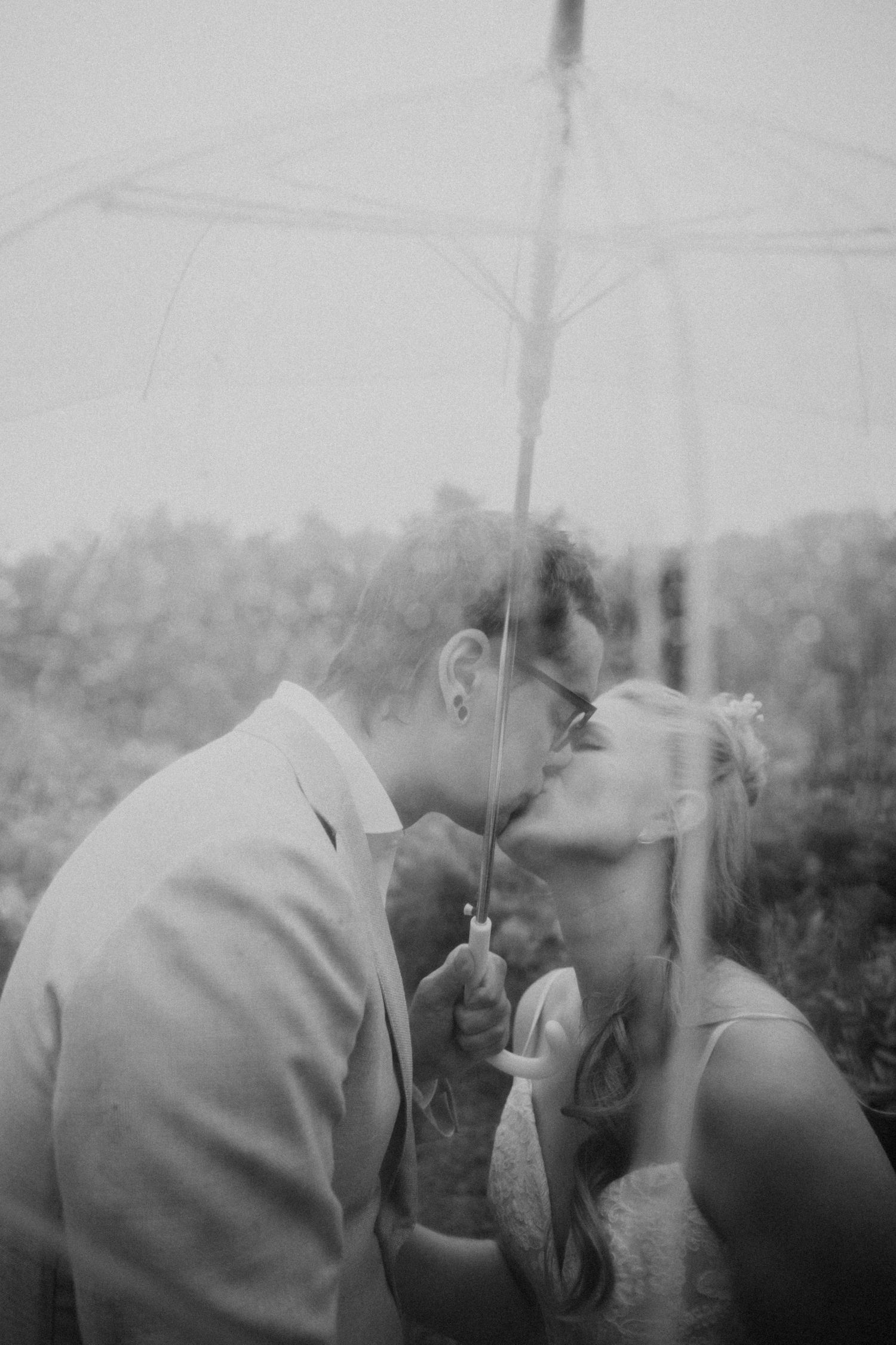 Bride and groom share a romantic kiss under a clear umbrella in the rain, black and white.
