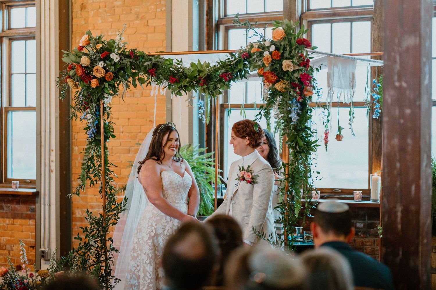 Two brides exchange vows under lush floral arch at sunlit industrial wedding venue.
