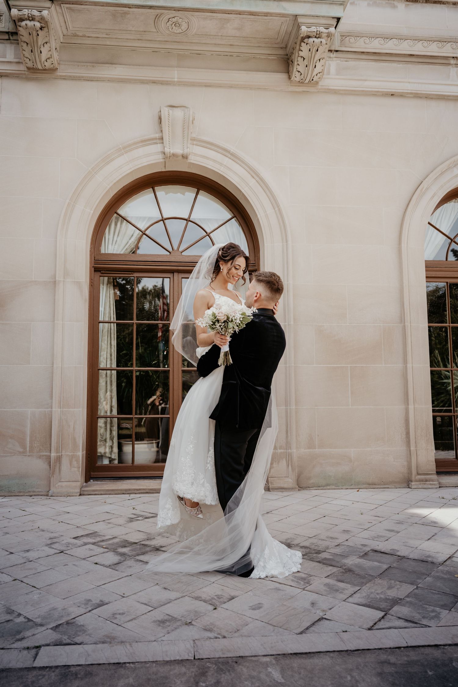 Bride and groom sharing romantic embrace before arched windows of elegant stone venue.