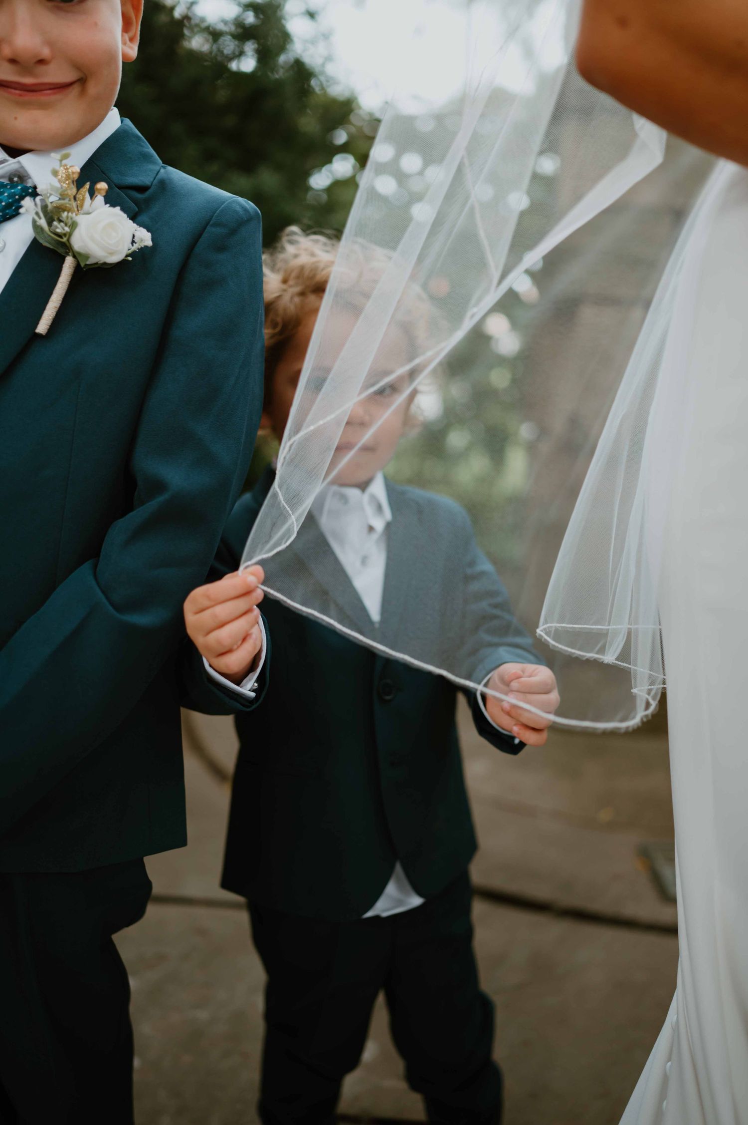 Young ring bearer playfully lifts bride's veil while standing next to older boy in teal suit at outdoor wedding.