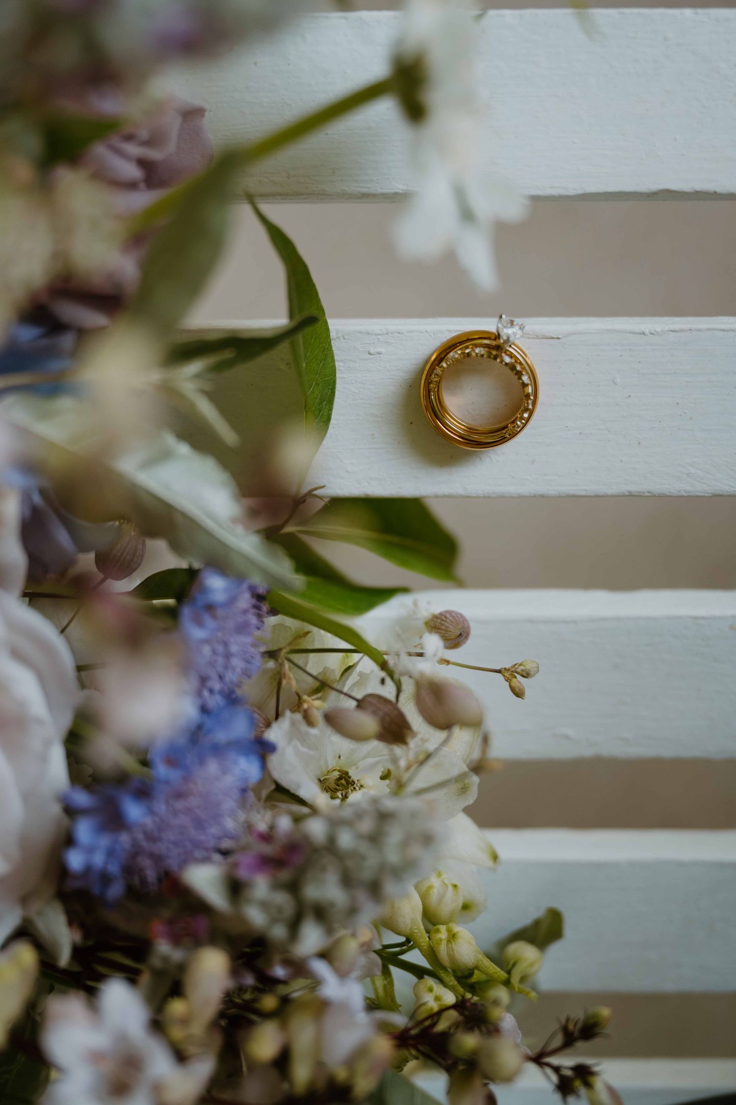 Two gold wedding rings displayed on white wooden shutters beside a soft floral bouquet.