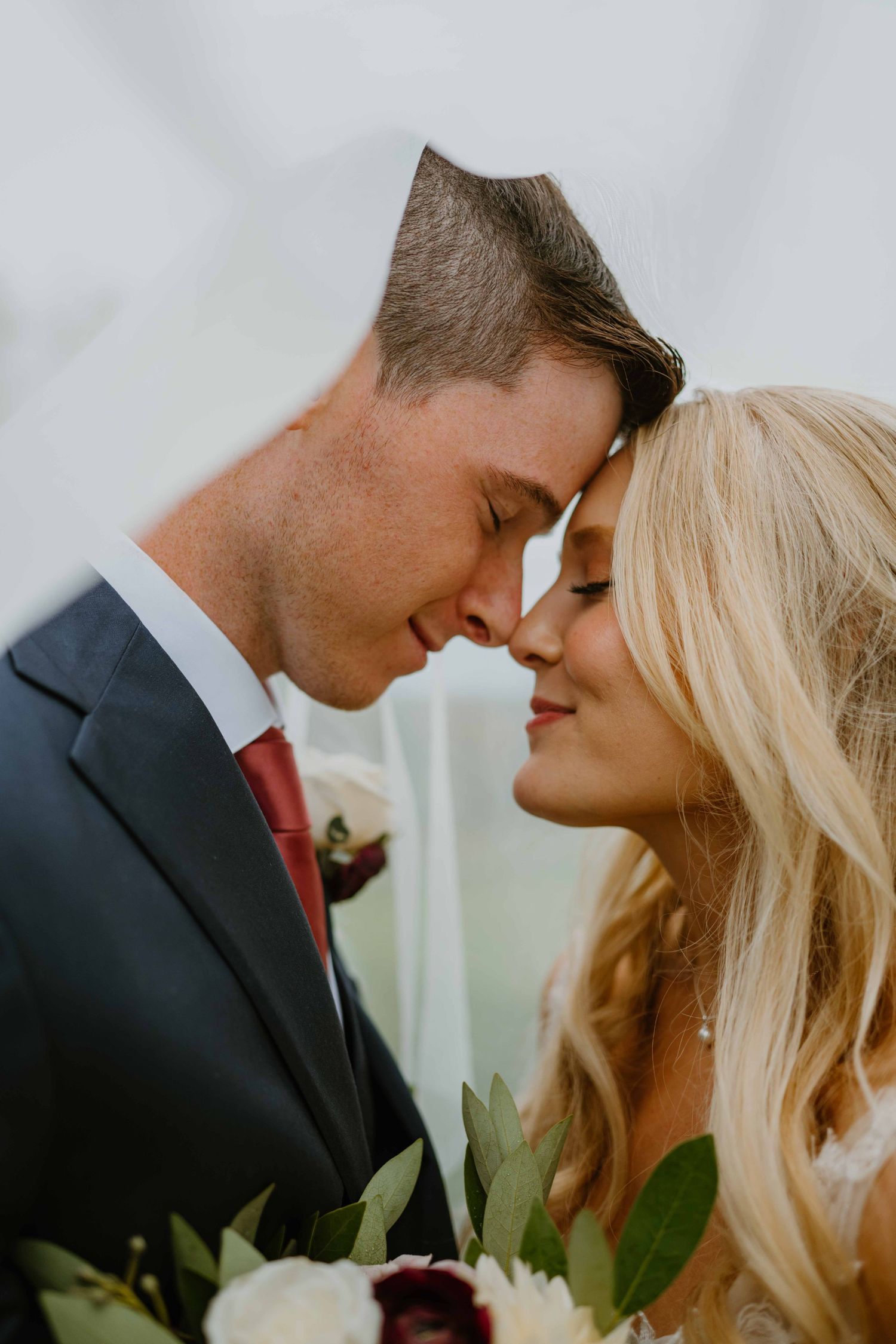 Bride and groom touching foreheads romantically holding burgundy wedding bouquet.