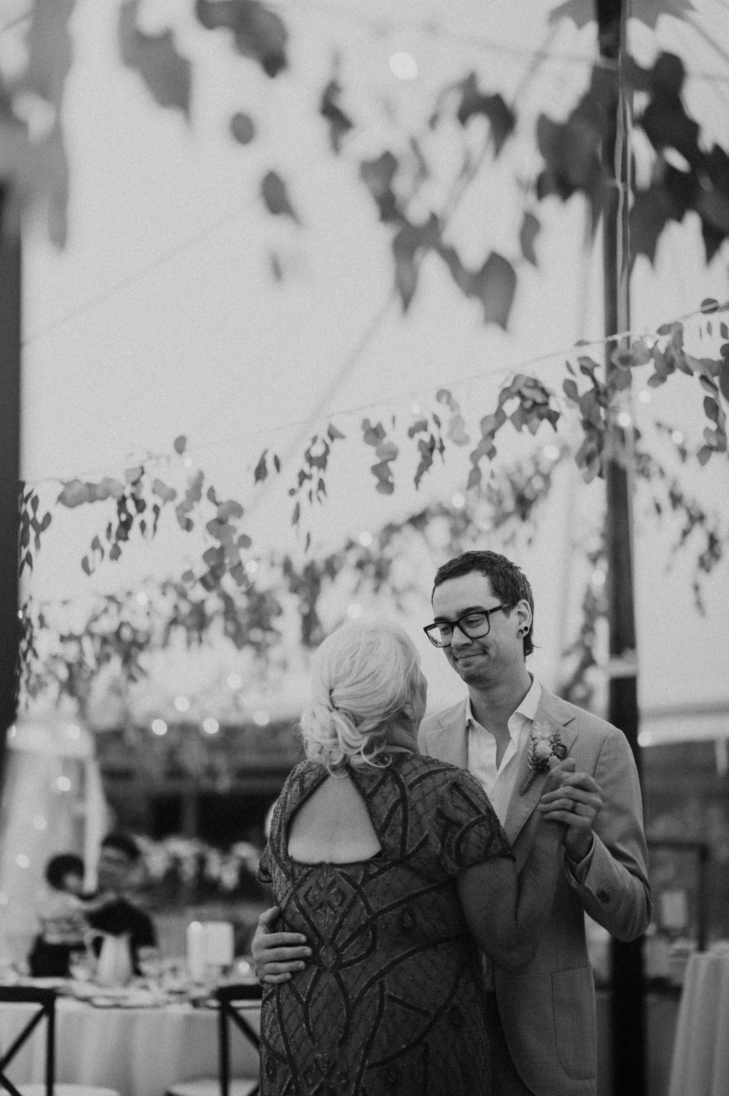 Groom in suit dancing with older woman under floral canopy at wedding reception in black and white.