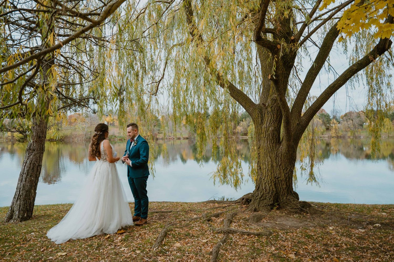 Bride and groom share intimate moment under willow tree by autumn lake during wedding photos.