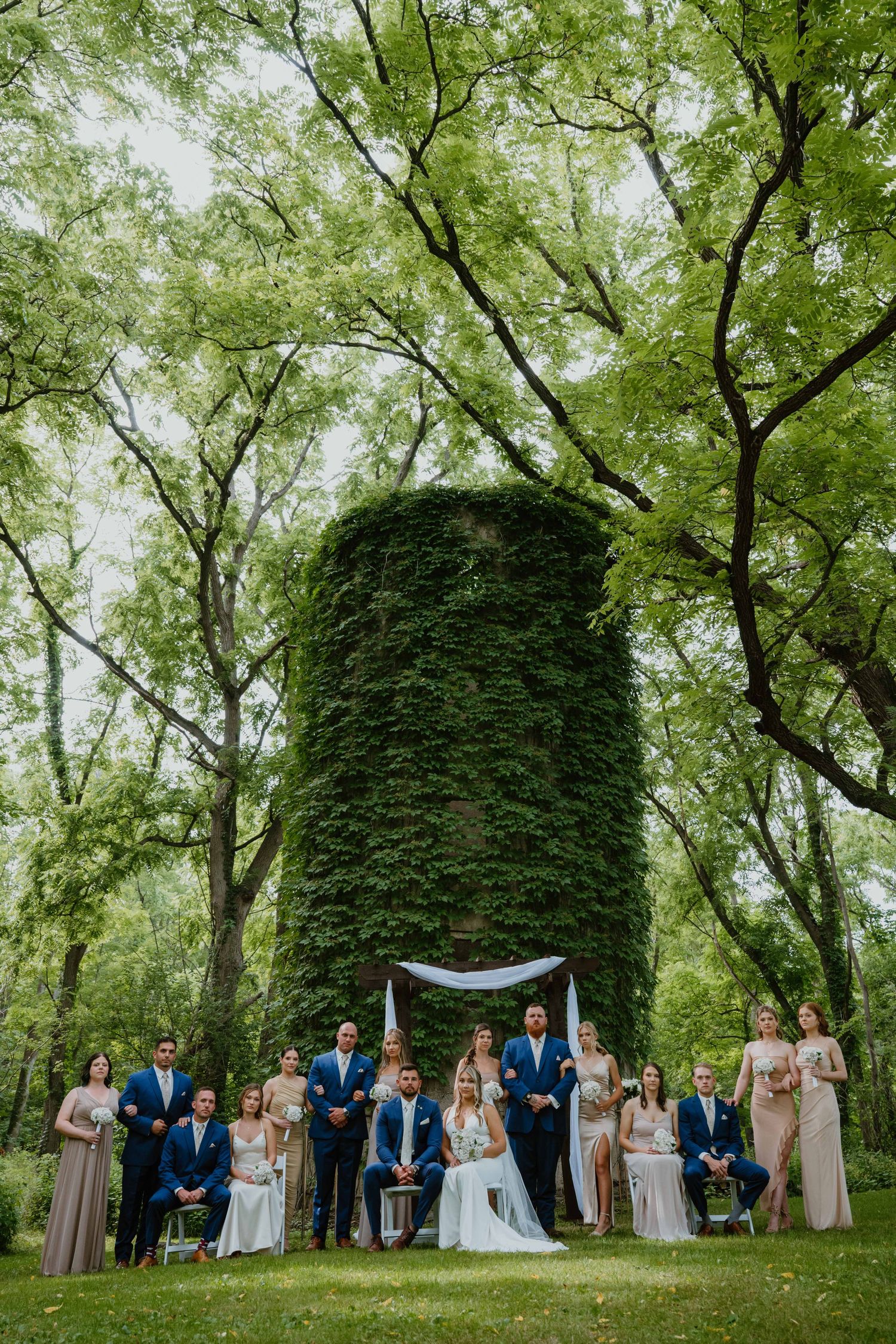 Wedding party poses before an ivy-covered tower surrounded by lush green trees.