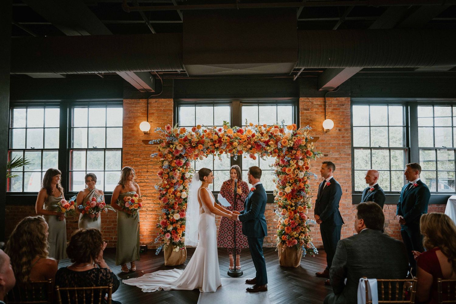 Wedding ceremony under colorful floral arch in rustic brick venue.