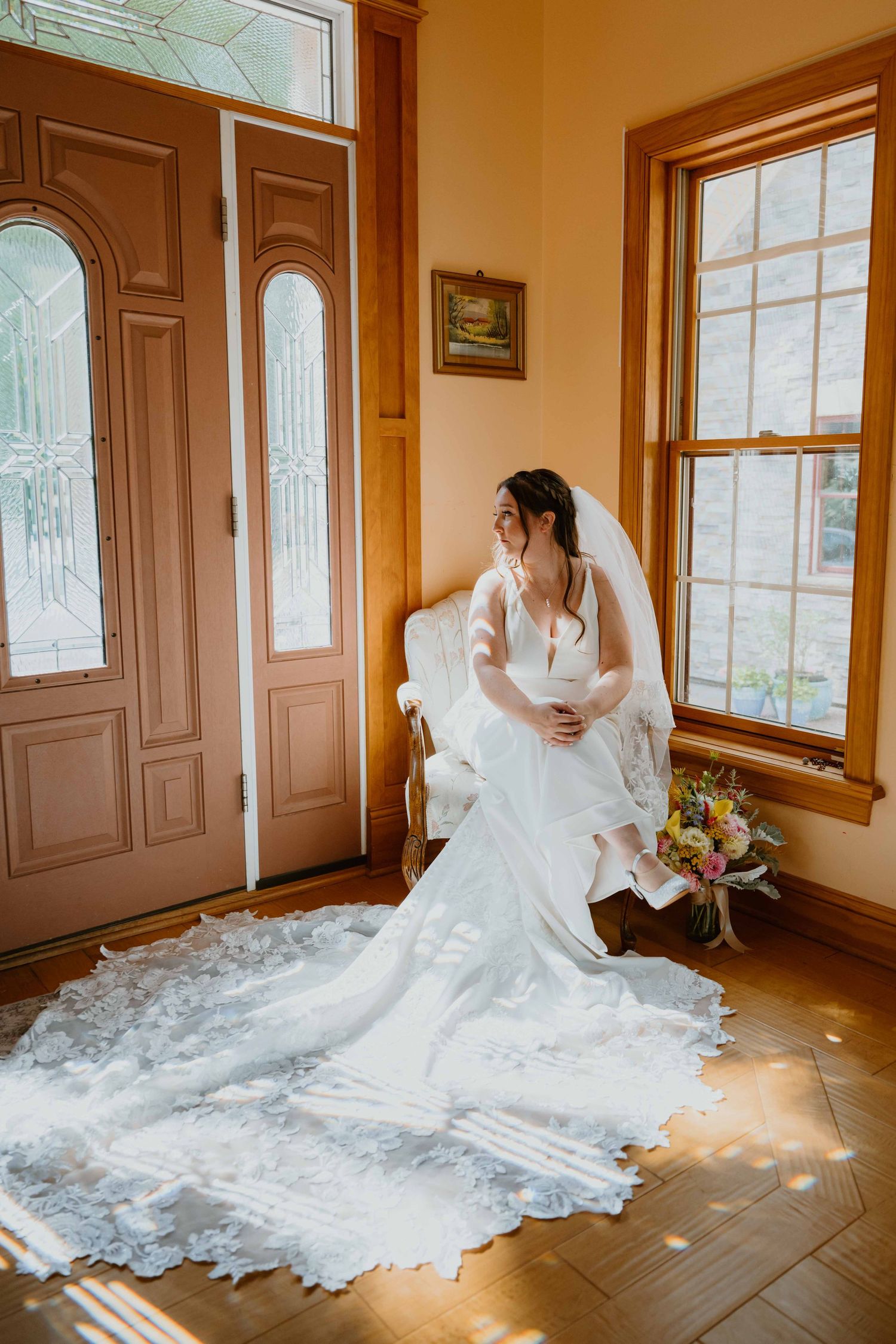 Bride in lace gown with cathedral veil sits by sunlit windows holding a bouquet.