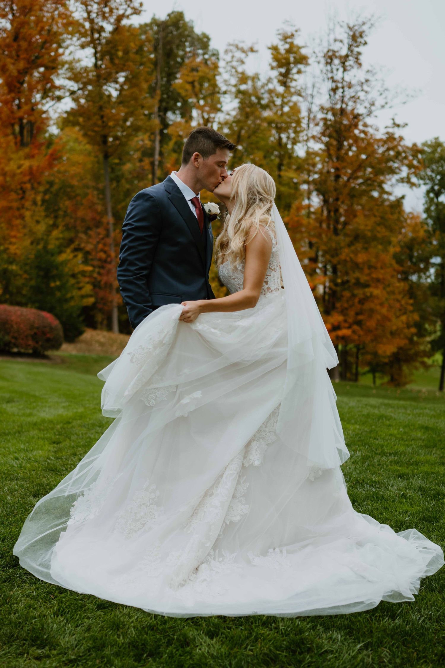 Bride and groom kissing on lush lawn surrounded by colorful autumn trees.