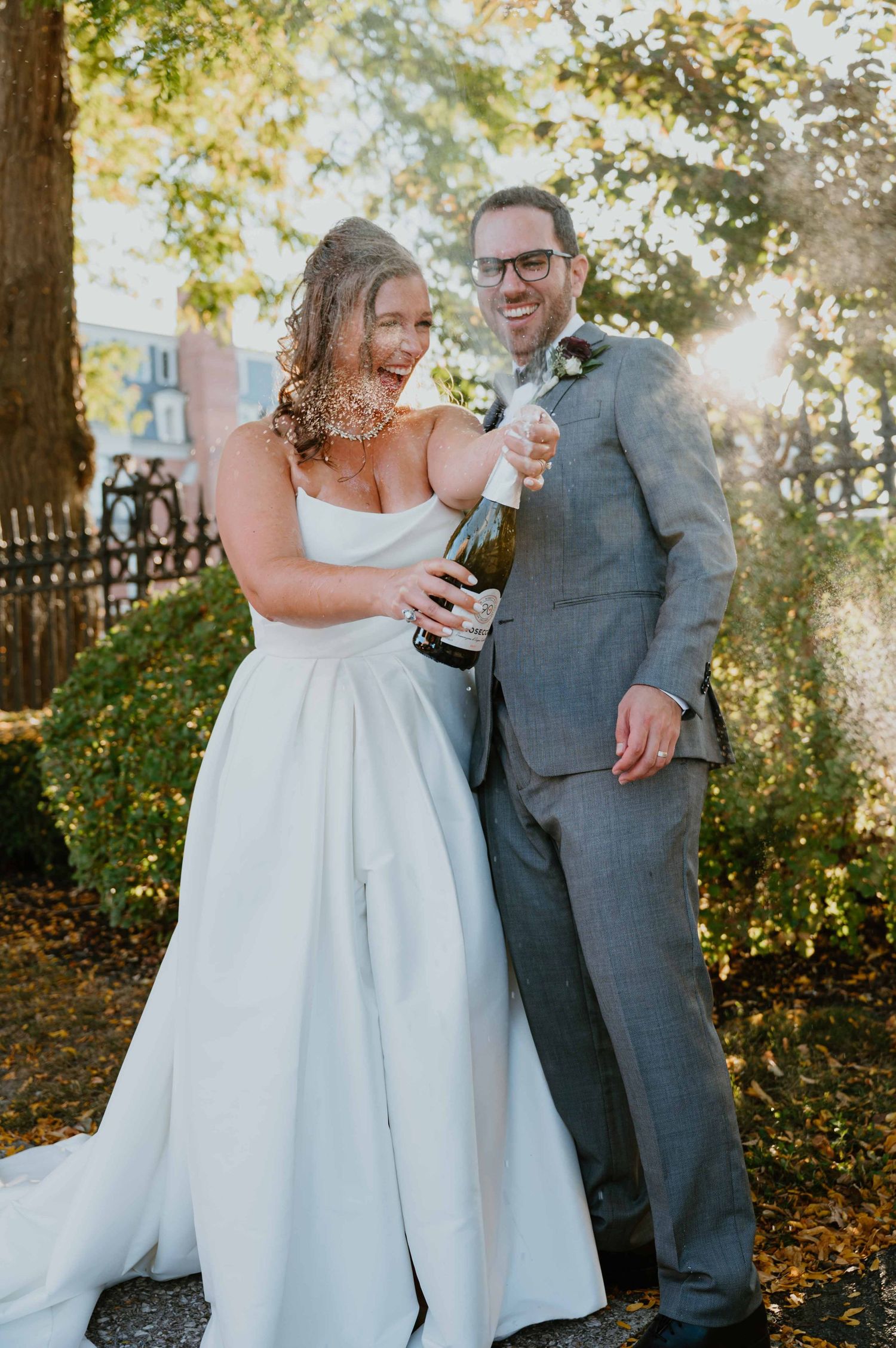 Joyful bride and groom pop champagne bottle outdoors in golden sunlight after their wedding ceremony.