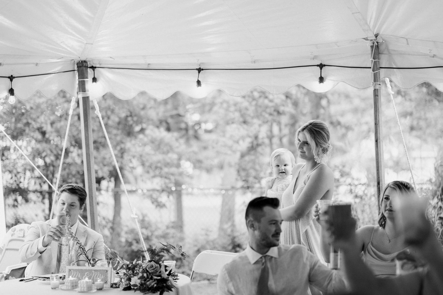 Black and white photo of wedding guests gathered under a tent with string lights.