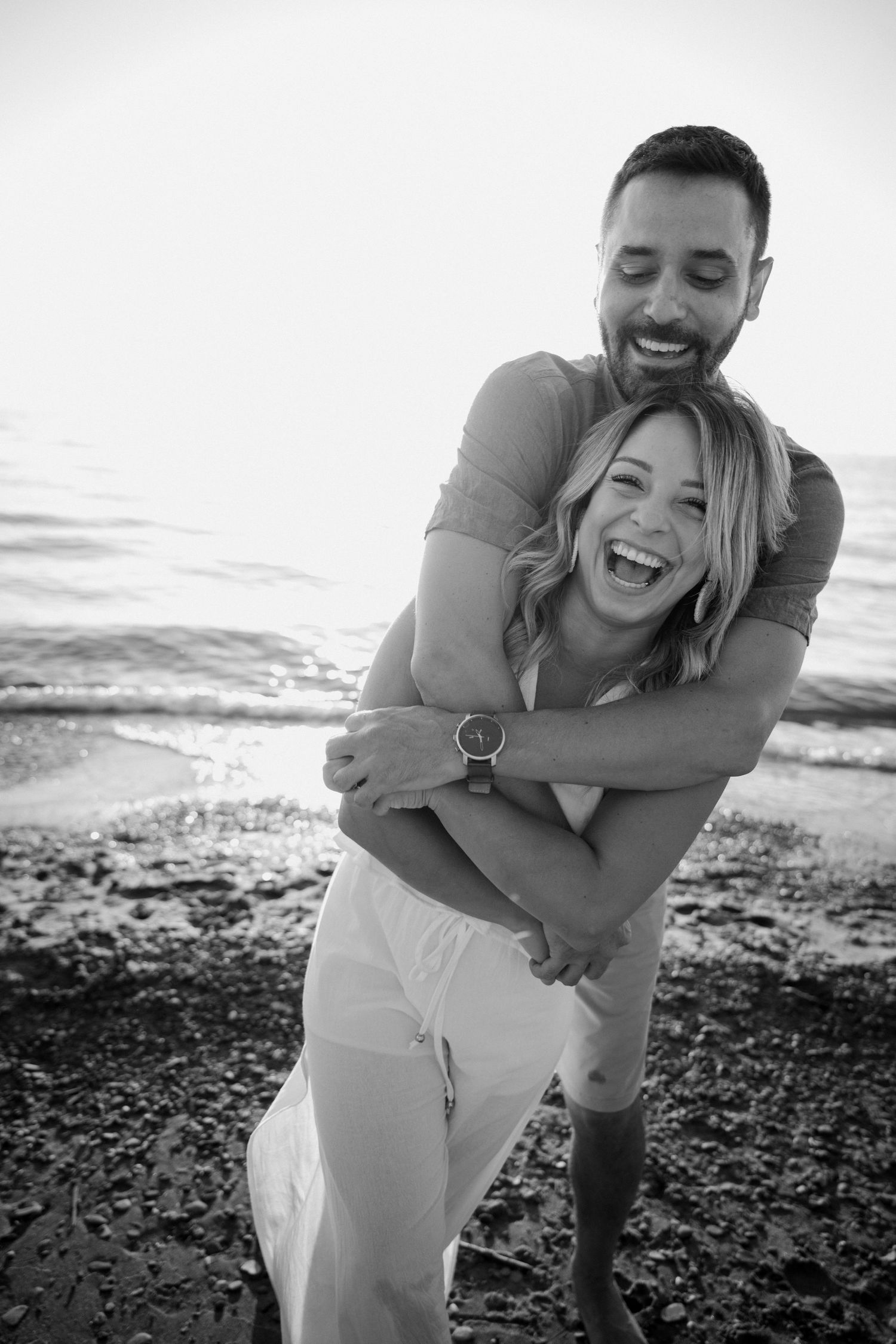 Black and white photo of joyful couple embracing and laughing together on a rocky shoreline.