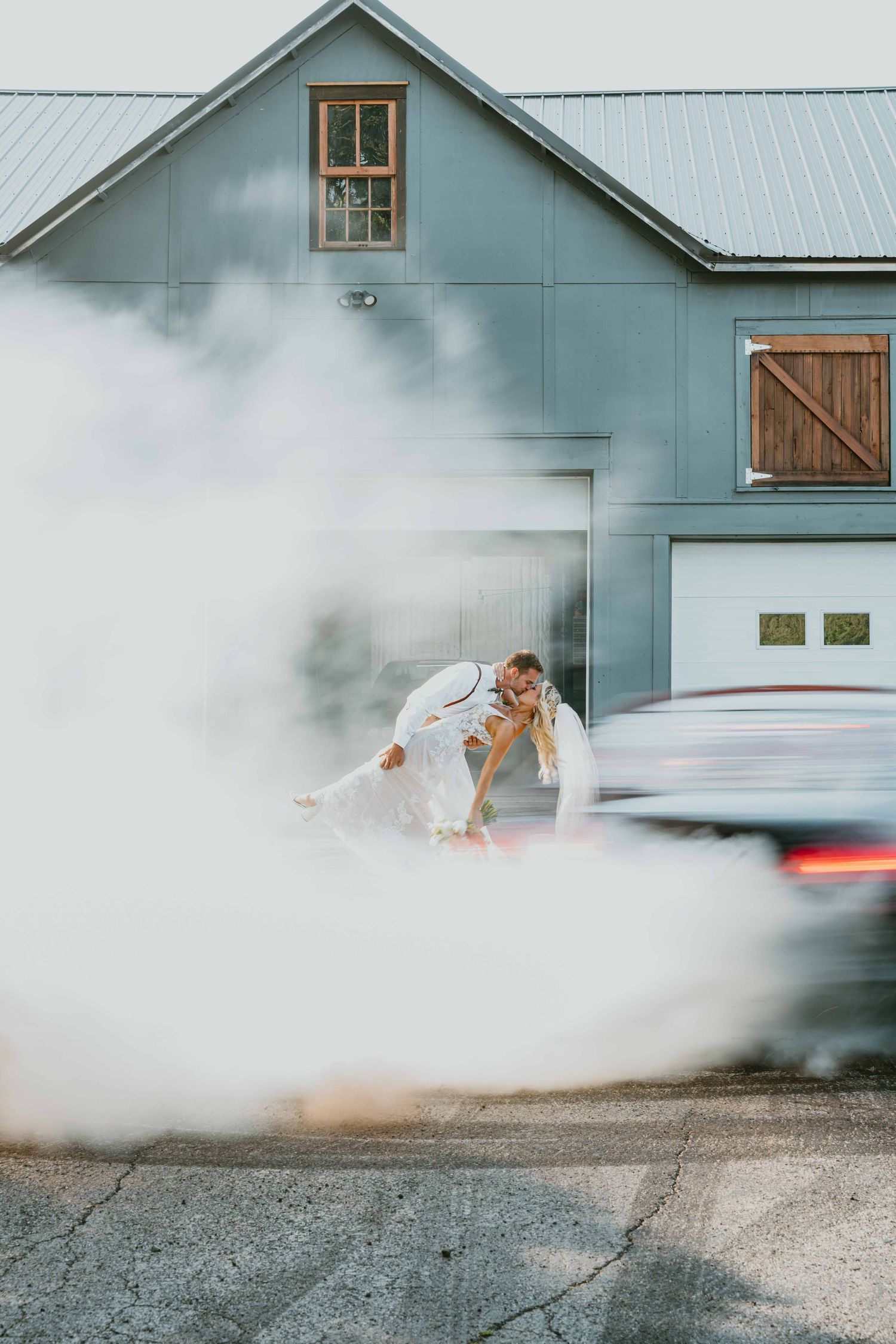 Bride and groom share a romantic dip kiss surrounded by dramatic smoke from a car burnout outside a blue barn.
