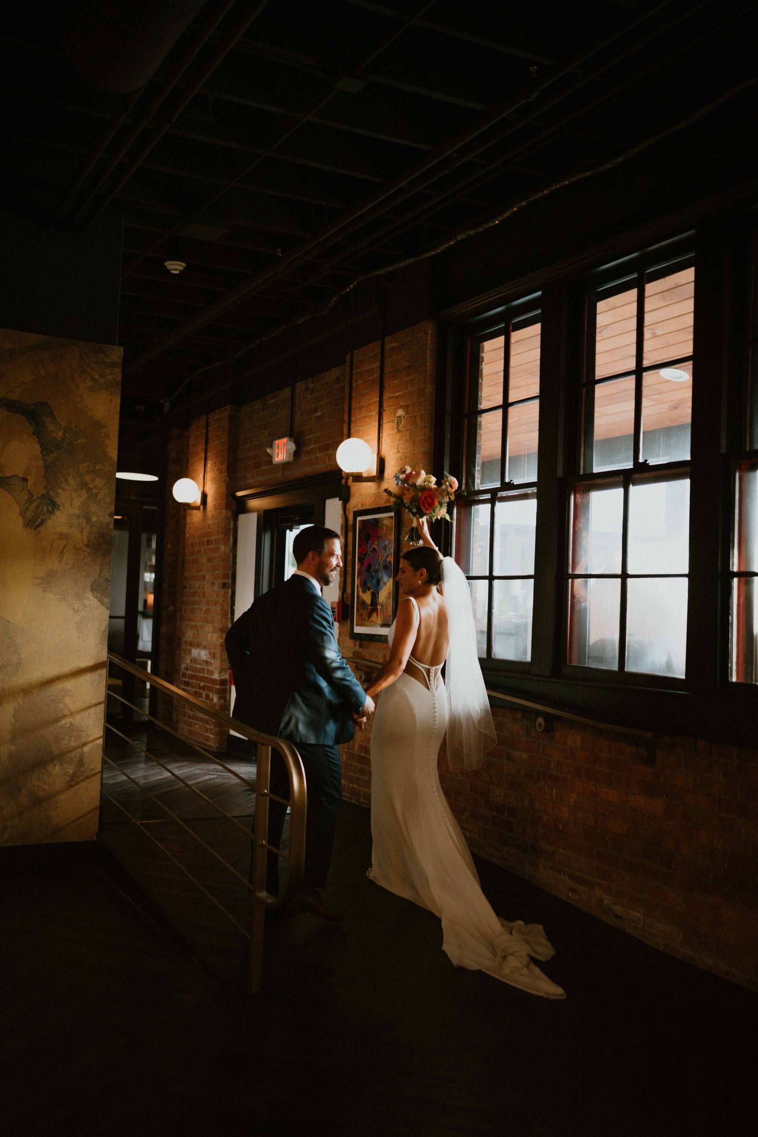 Bride and groom share intimate moment by tall windows in dark venue.