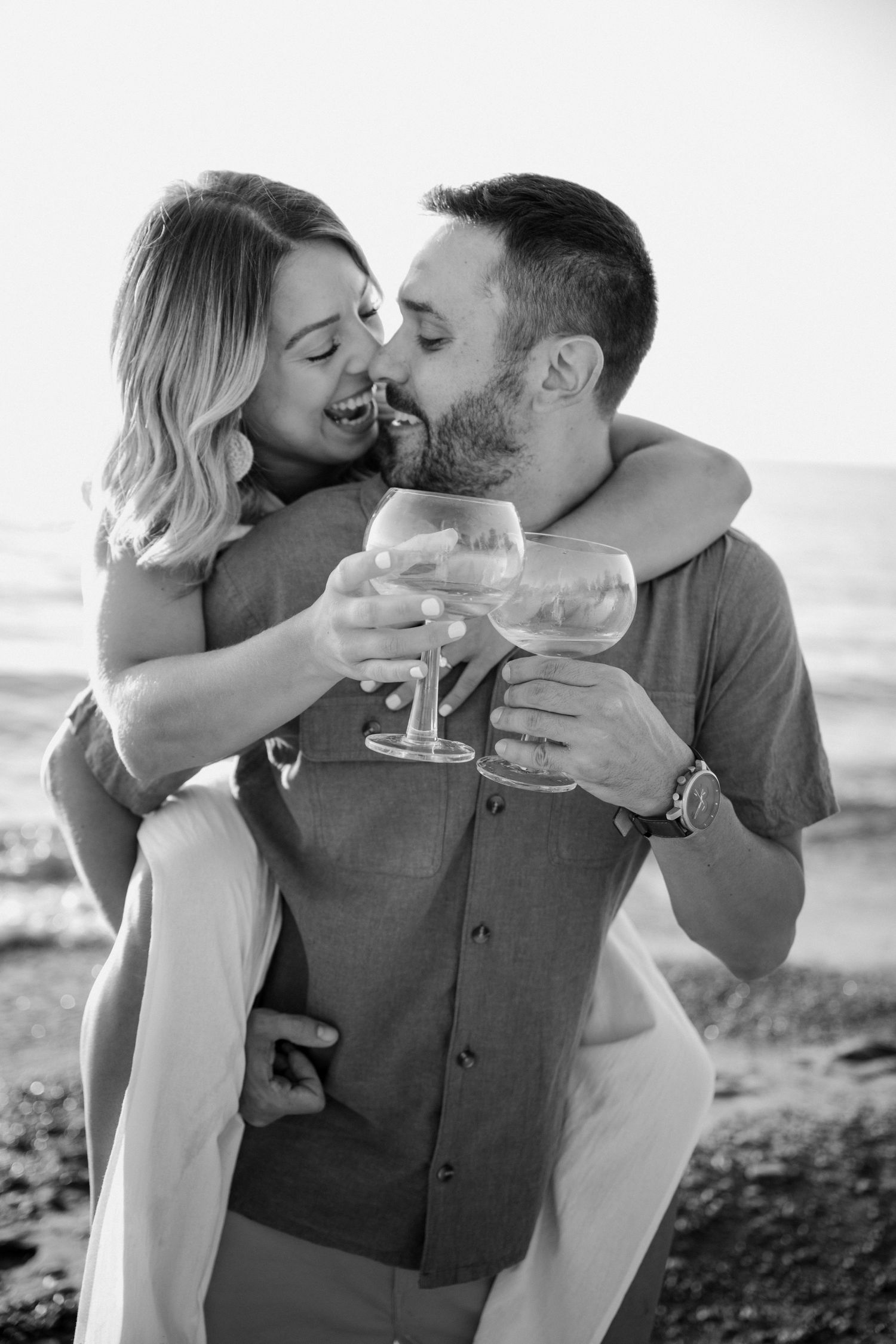 Black and white photo of laughing couple toasting with wine glasses during a romantic beach engagement session.