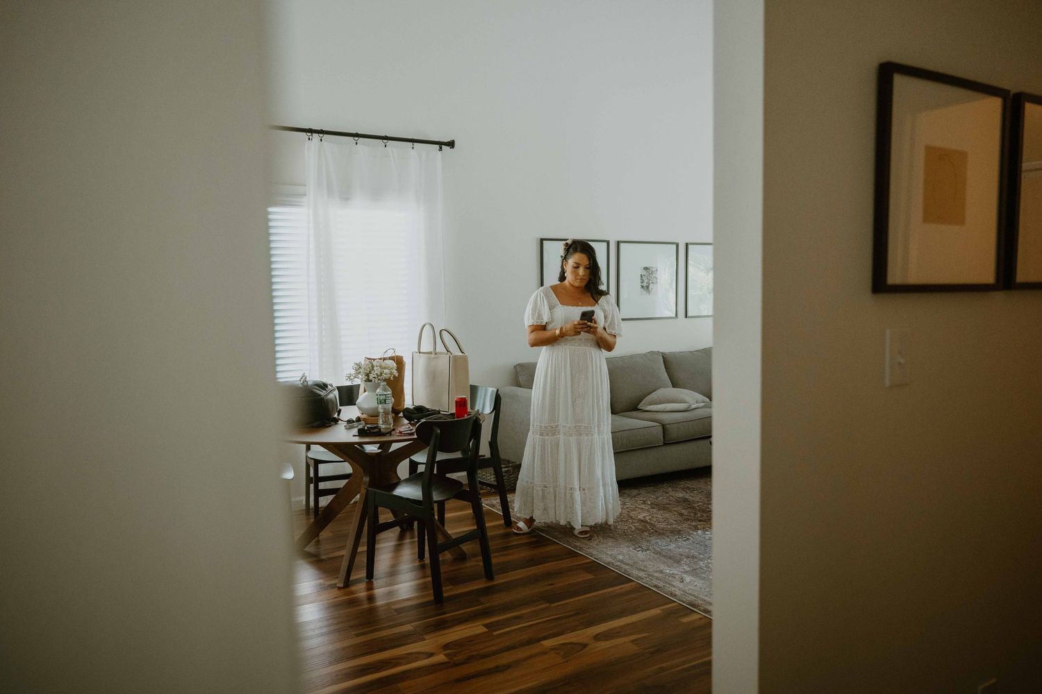 Bride in white dress stands in a modern living room looking at her phone.