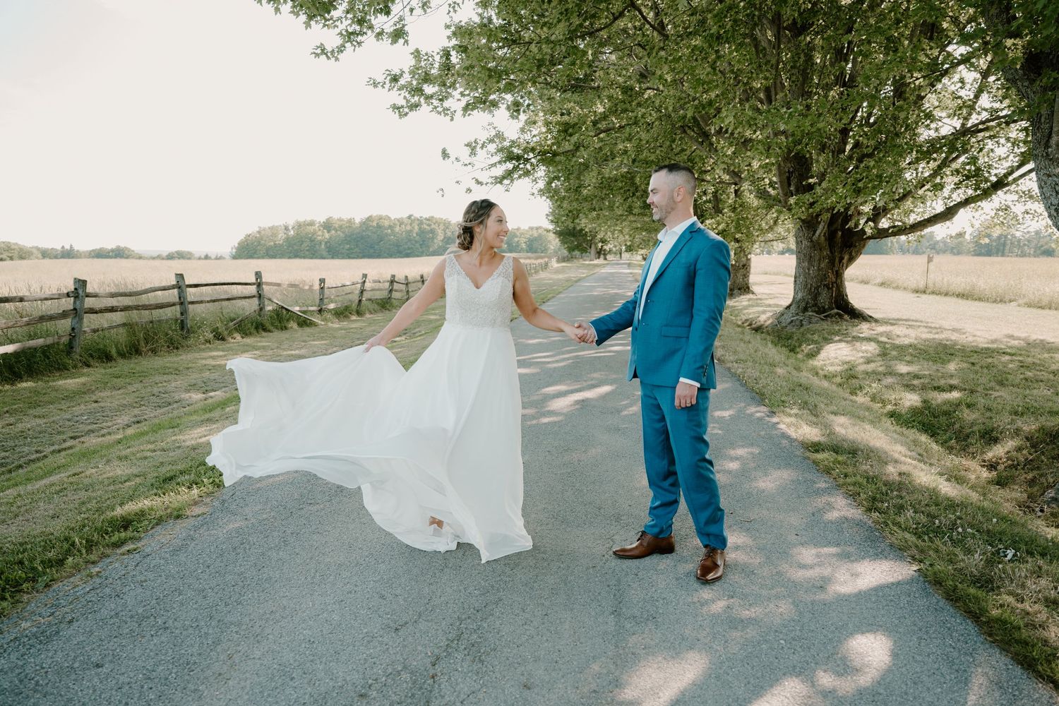 Bride and groom hold hands on scenic country road with flowing dress caught in breeze.