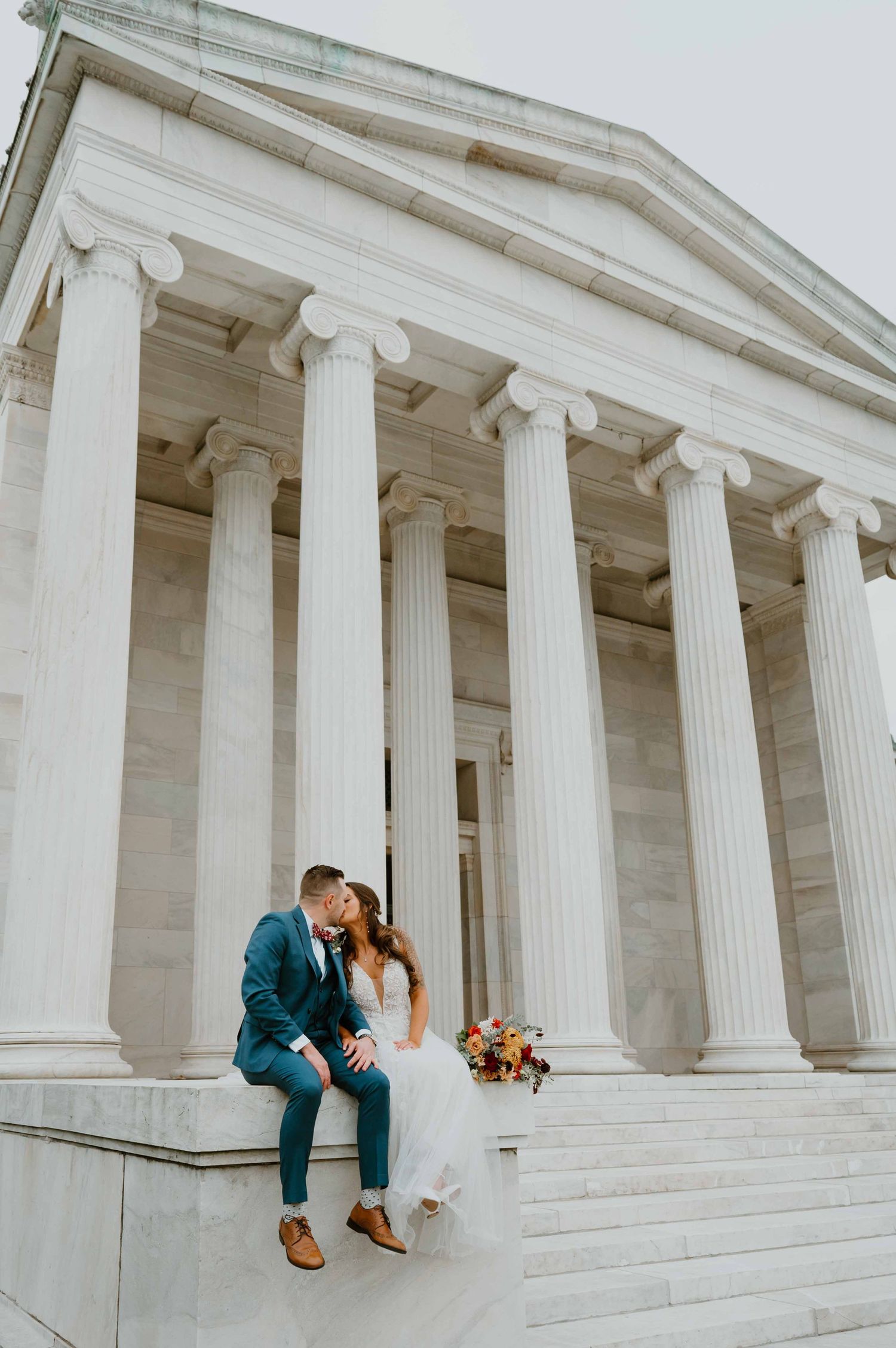 Bride and groom share a kiss on steps of grand classical columned building with fall bouquet.