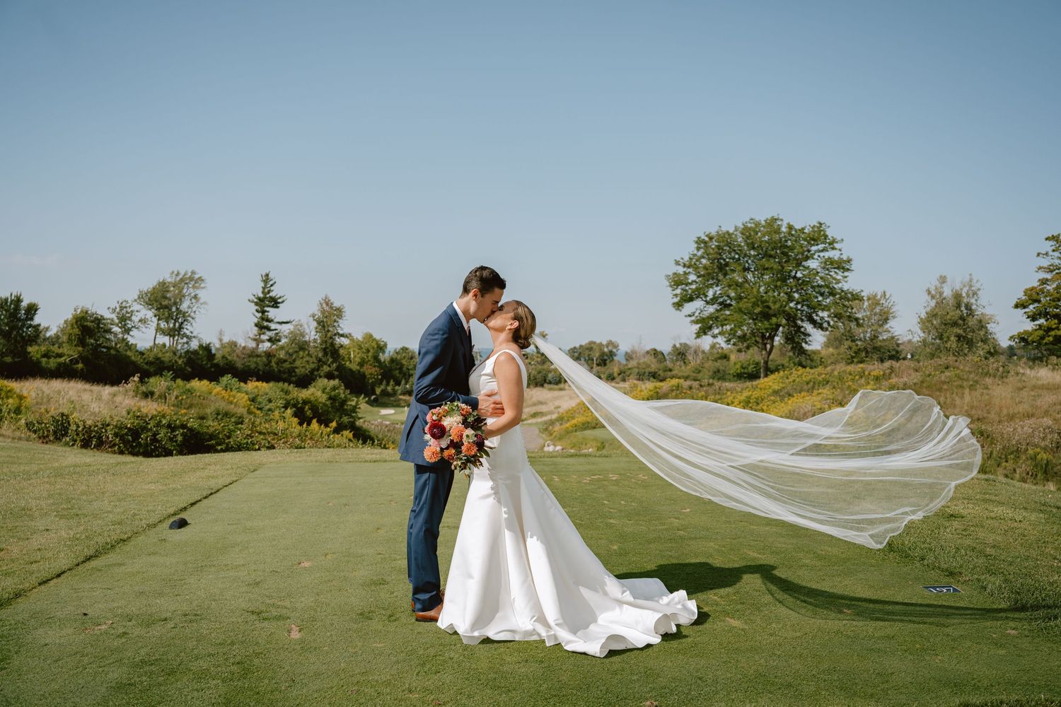 Bride and groom kissing on a golf course with the bride's long veil flowing in the breeze.