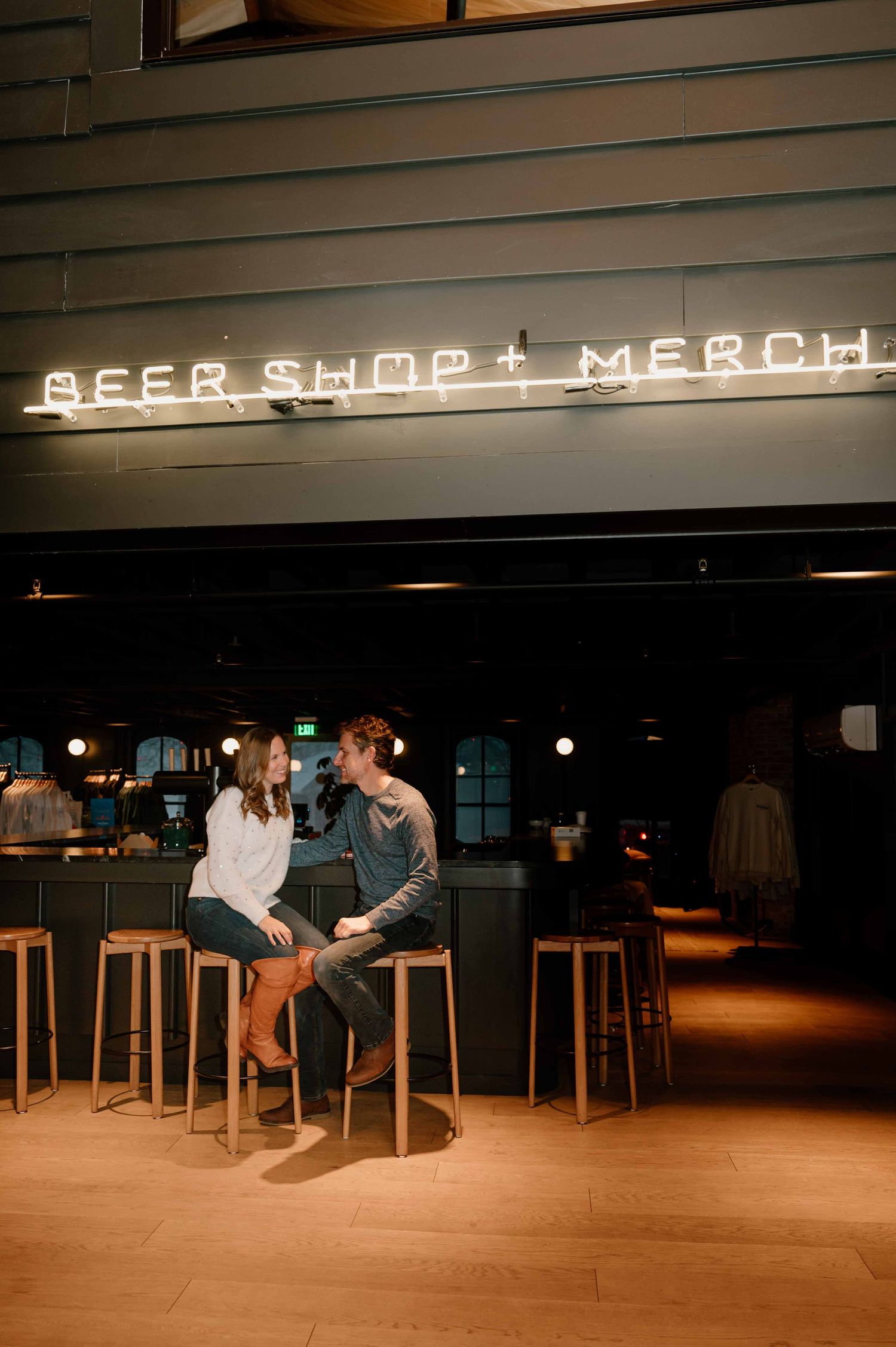 Couple sitting on barstools facing each other under neon Beer Shop and Merch sign at brewery.