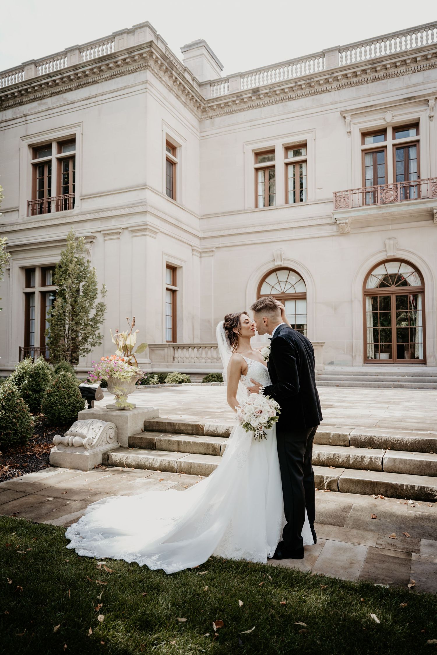 Bride and groom kissing in garden of stunning white neoclassical mansion with steps.