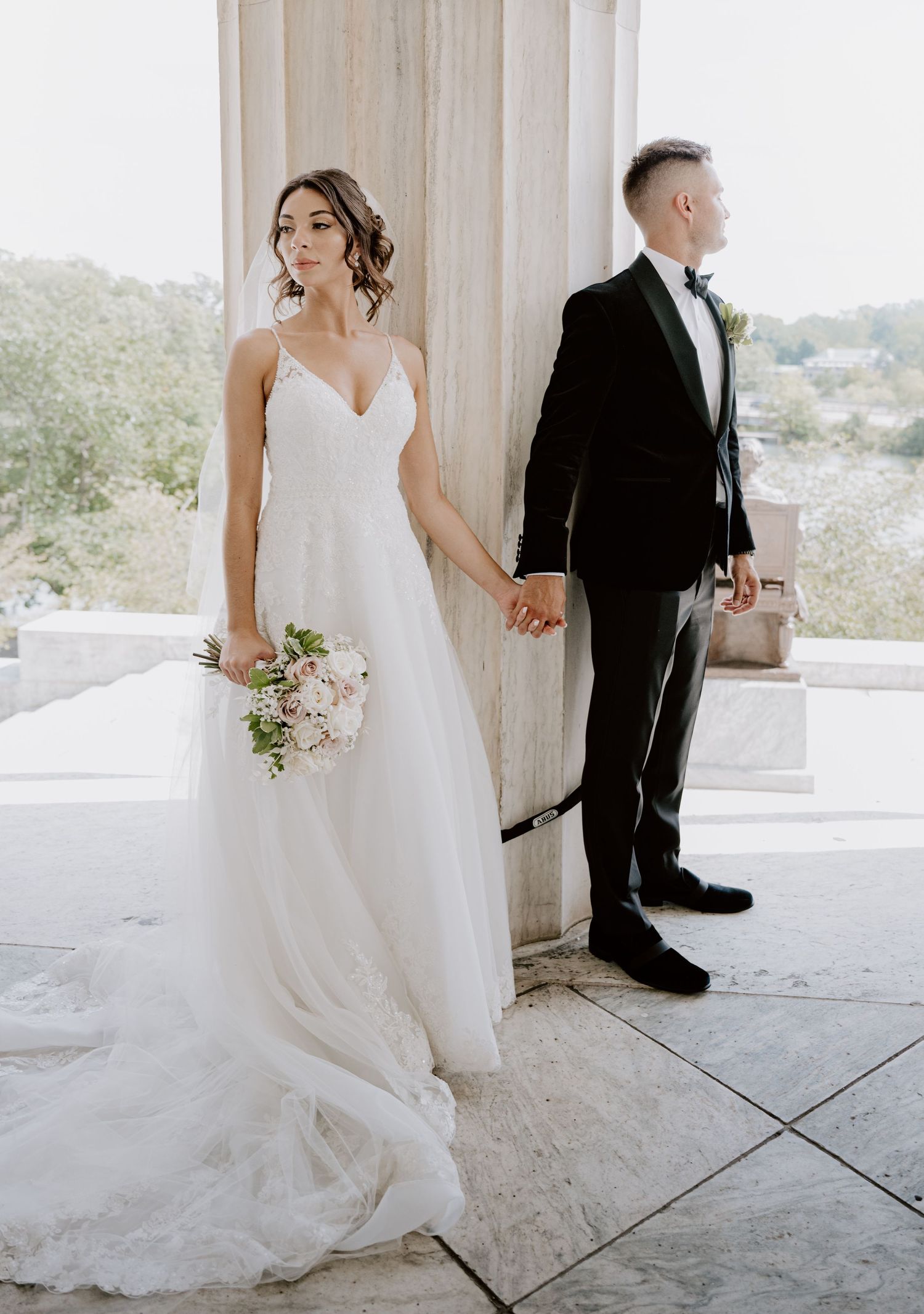 Bride holding pink bouquet and groom standing back-to-back between marble columns.