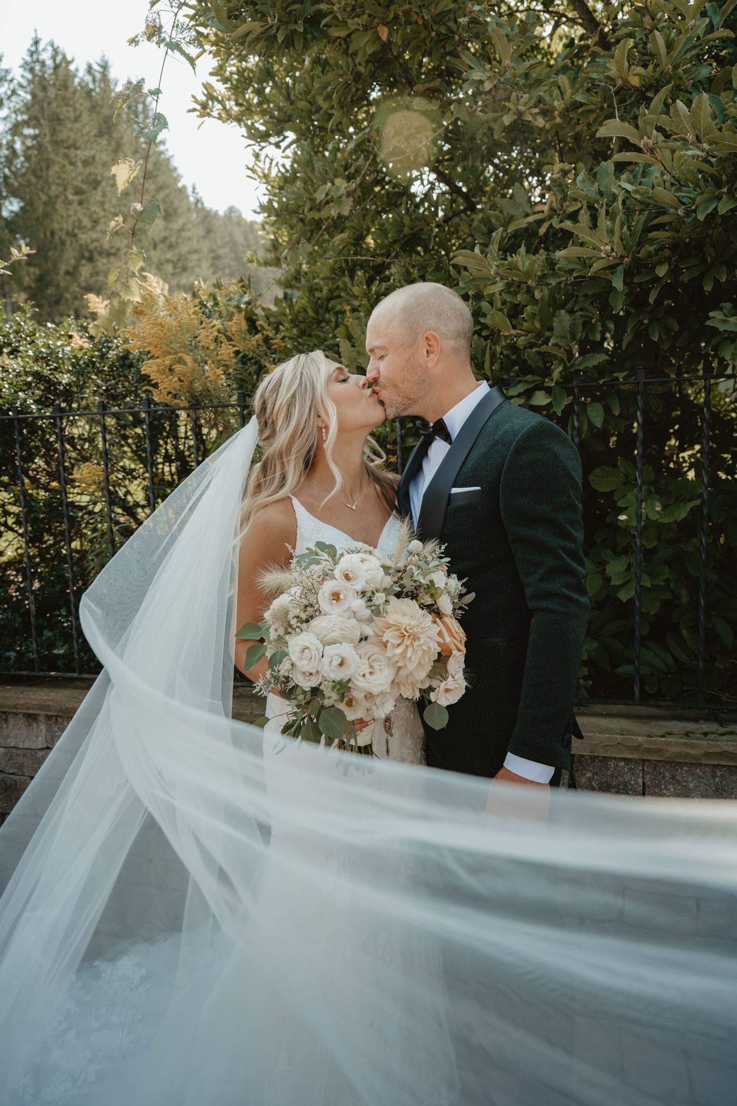 Bride and groom sharing a kiss, veil flowing in the wind, bride holding white floral bouquet.