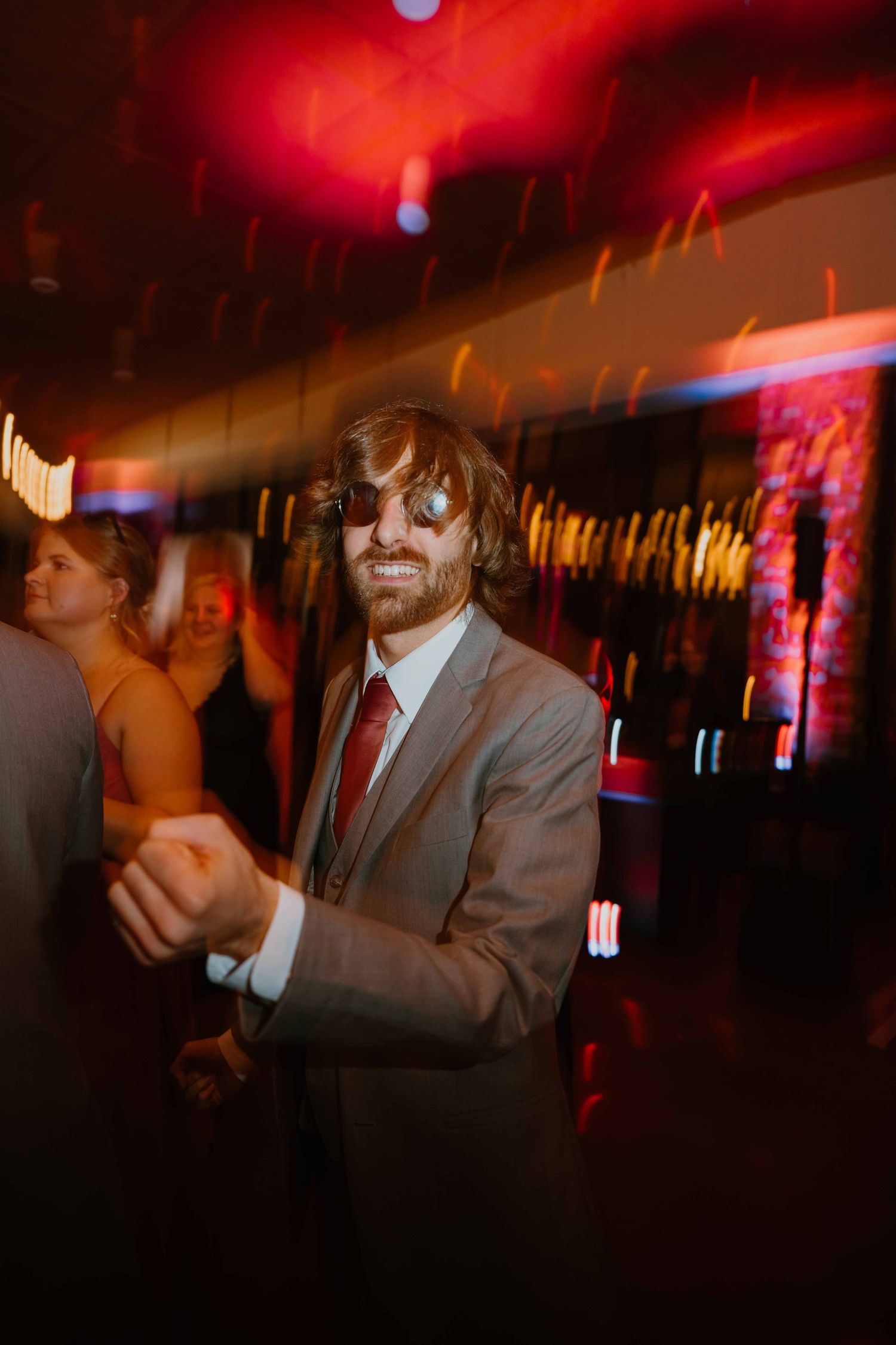 Smiling male wedding guest in gray suit and sunglasses dancing at a lively wedding reception.
