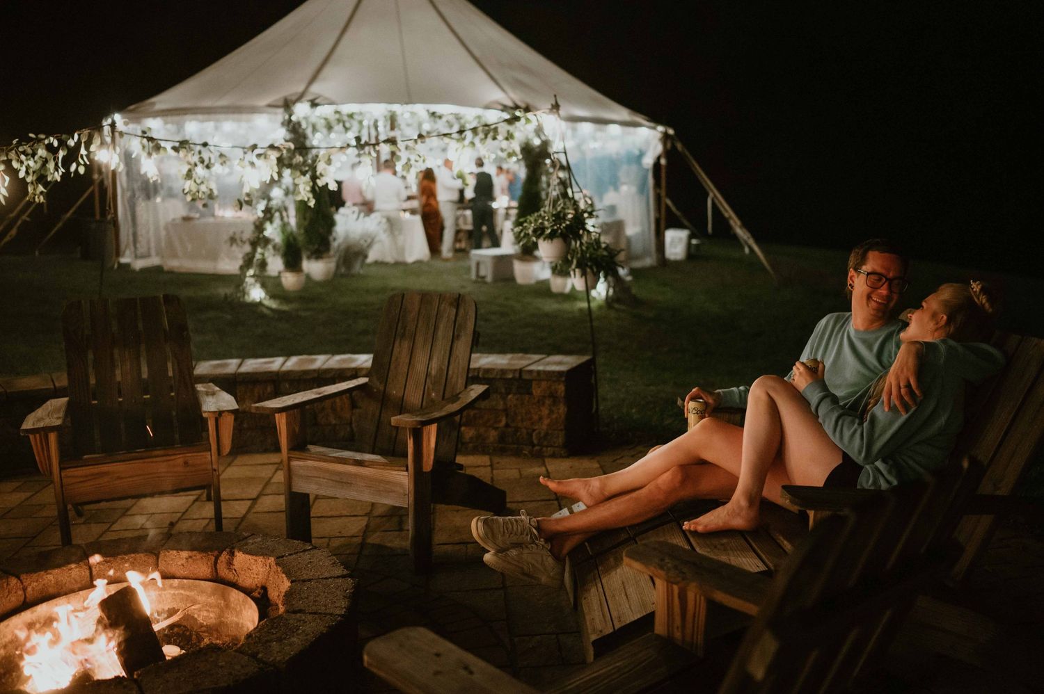 Couple relaxes by outdoor fire pit at night with illuminated wedding tent in background.