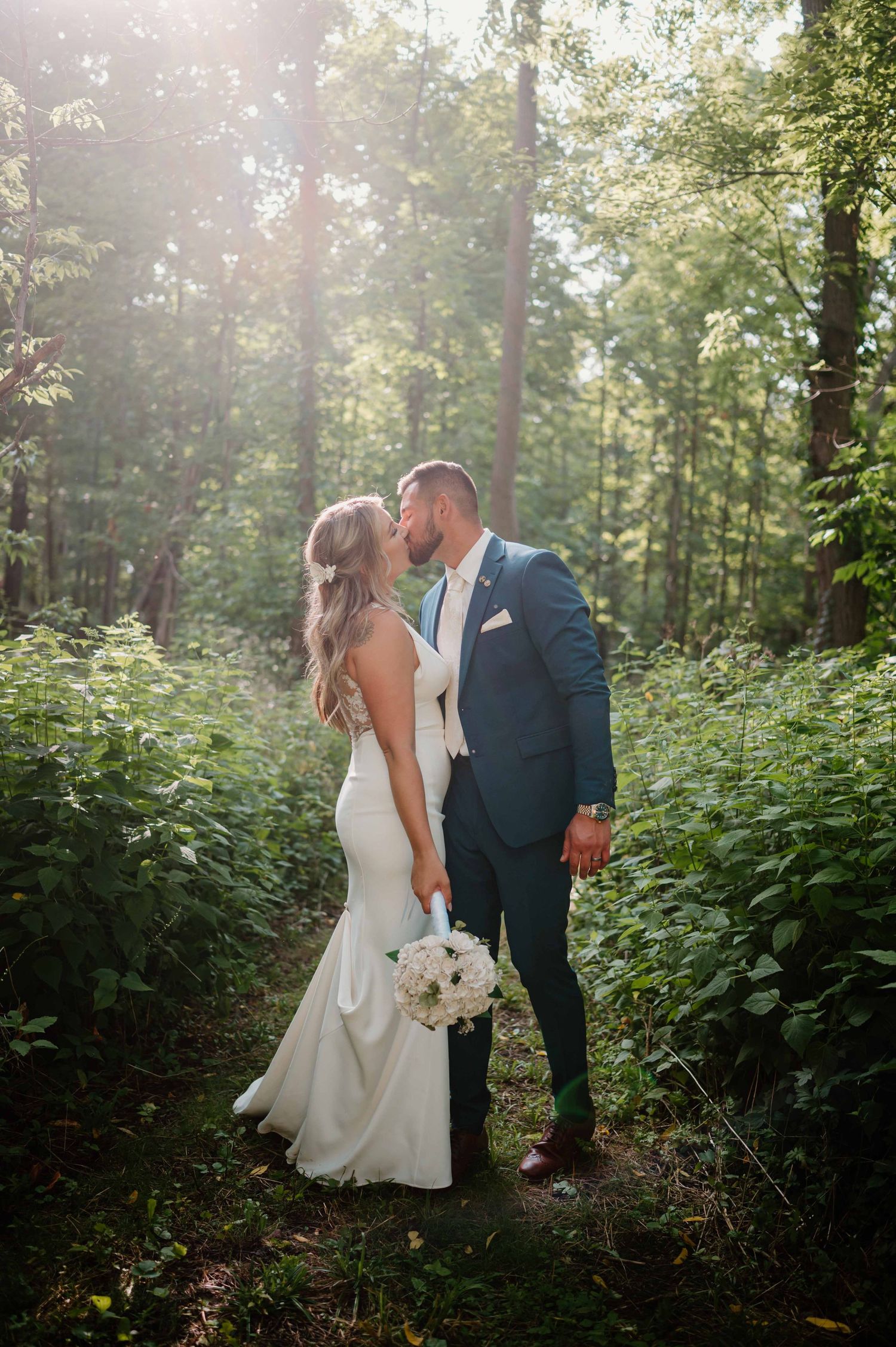 Bride and groom share a kiss on a sunlit forest path, bride holding a white bouquet.